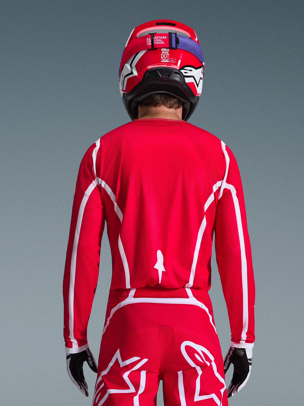 Vue arrière d'un pilote de motocross portant le maillot Alpinestars 2026 Fluid Apex en rouge et blanc, présentant des motifs géométriques blancs, assorti d'un pantalon de motocross et d'un casque rouge, équipement de course complet pour la performance tout-terrain.