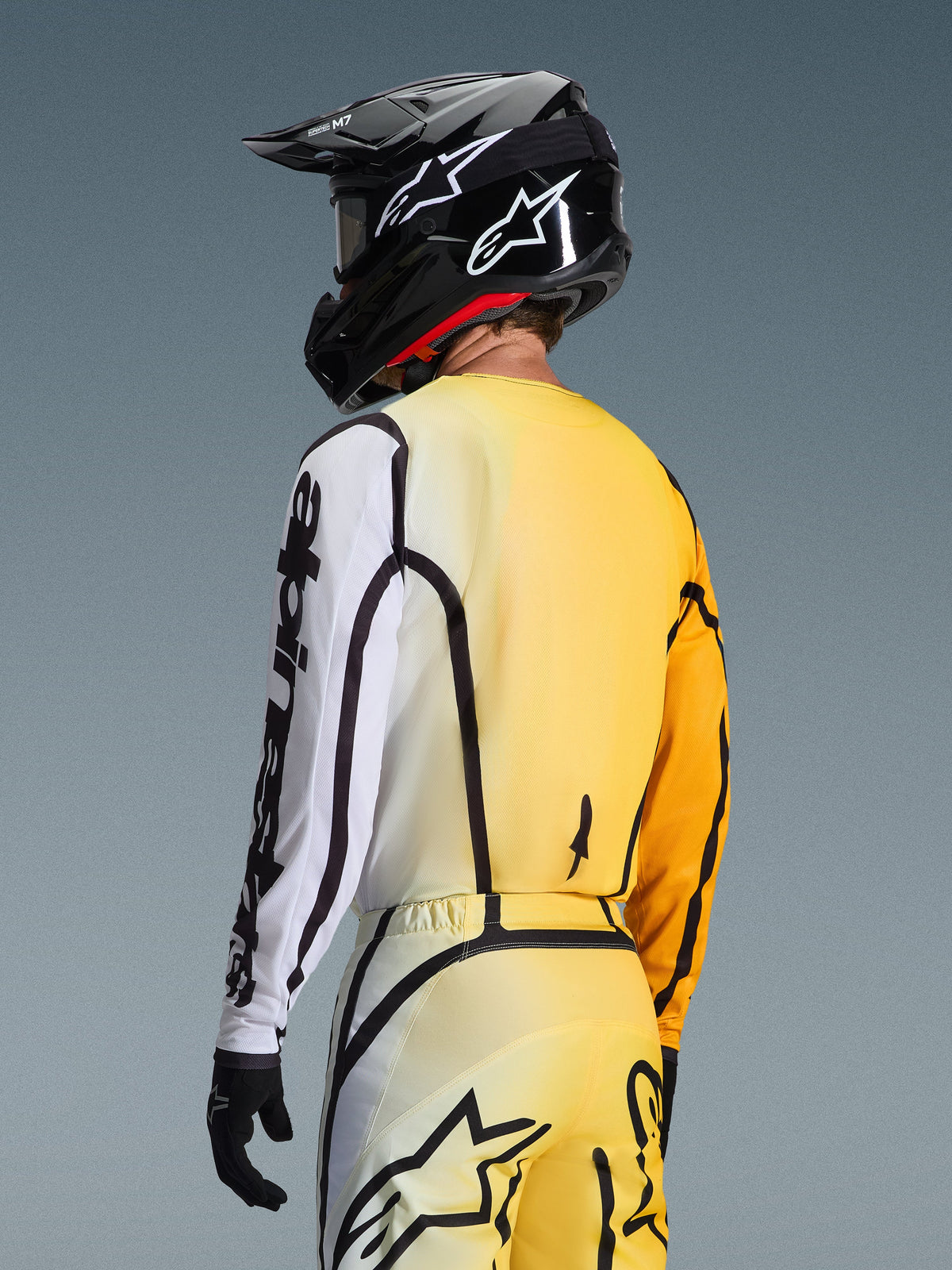 Vue de trois-quarts arrière d'un pilote de motocross portant le maillot Alpinestars Fluid Apex 2026 en Jaune Noir, avec un dégradé du jaune au blanc, logo Alpinestars noir sur la manche, pantalon assorti et casque de motocross noir.