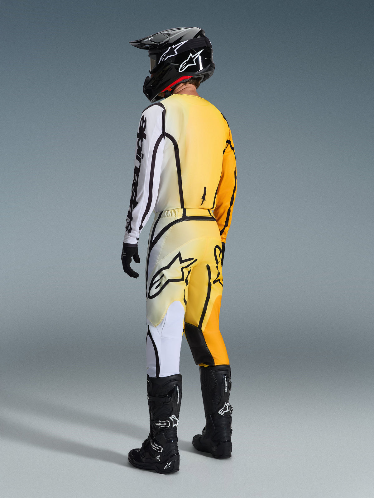 Pilote de motocross vu de dos portant le maillot Alpinestars Fluid Apex 2026 et le pantalon assorti, tenue Jaune Noir avec dégradés du jaune au blanc et lignes graphiques noires marquées, accompagnée d'un casque noir, de gants noirs et blancs et de bottes de motocross noires