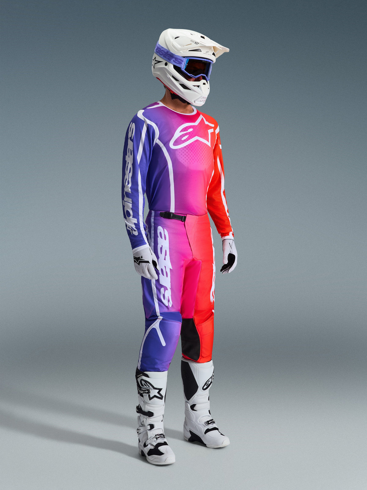 Pilote de motocross portant un équipement de course complet, maillot et pantalon Fluid Apex 2026 avec un dégradé éclatant de violet, rose et orange, casque blanc avec lunettes bleues, bottes blanches et noires, et gants blancs, tenue coordonnée pour le motocross tout-terrain.