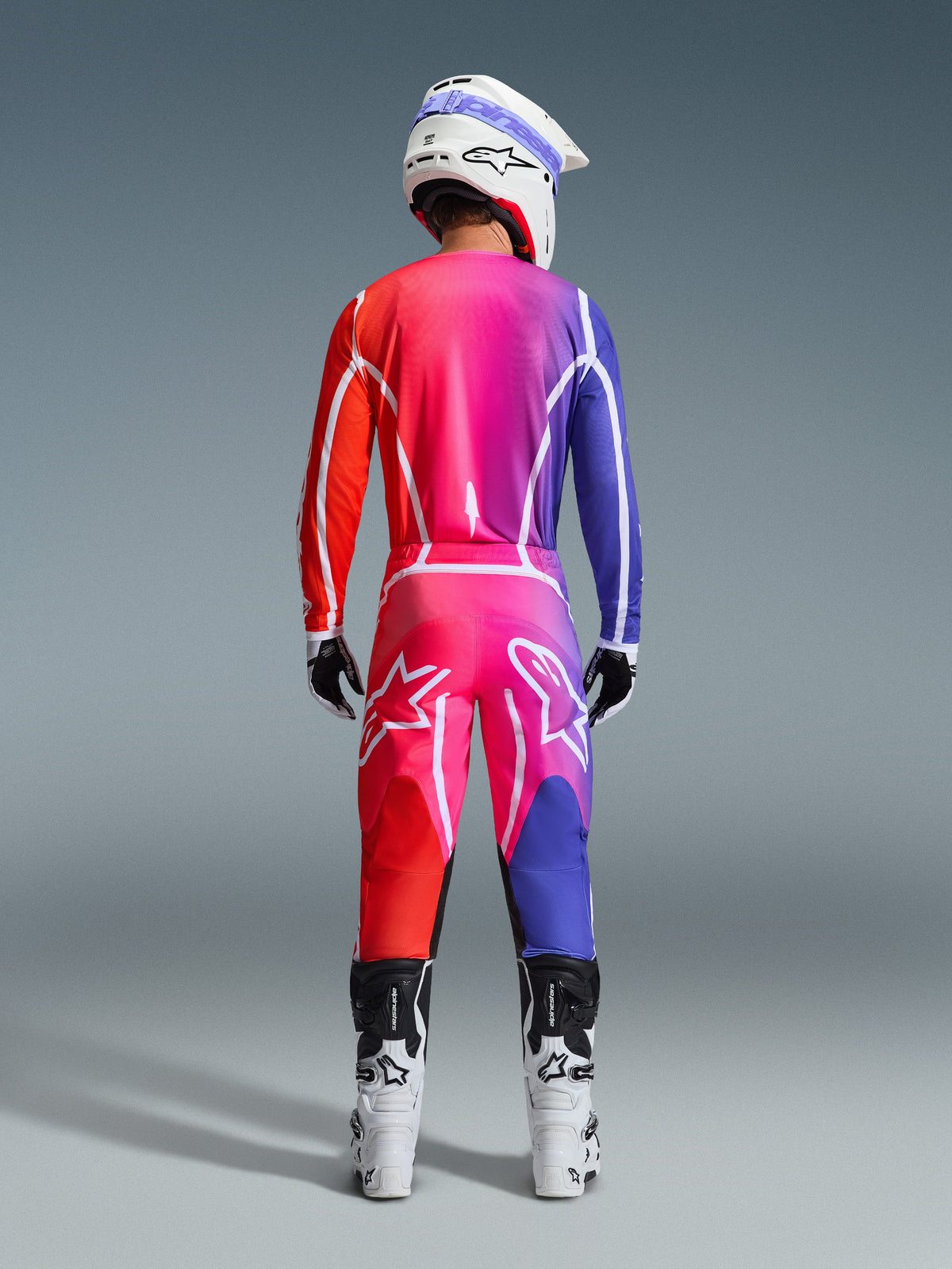 Alpinestars 2026 Fluid Apex Gear Set, Rückansicht eines Motocross-Fahrers in Jersey und Hose mit leuchtendem Farbverlauf in Rot, Pink und Violett, weiße Grafiklinien und große Stern-Logos, ergänzt durch einen weißen Helm und weiße Stiefel.