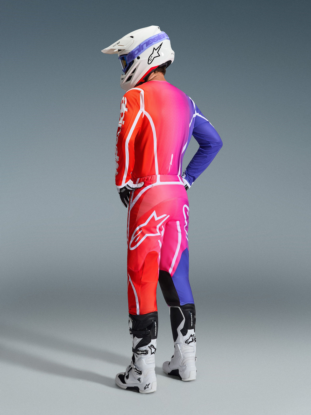Motocross-Fahrer im Alpinestars 2026 Fluid Apex Gear Set, lebendiger Farbverlauf von Orange zu Pink und Lila mit markantem weißen Branding und Stern-Logos, kombiniert mit einem weißen Helm, einer blauen Brille und weißen Stiefeln, Studioaufnahme aus einer rückwärtigen Drei-Viertel-Perspektive.