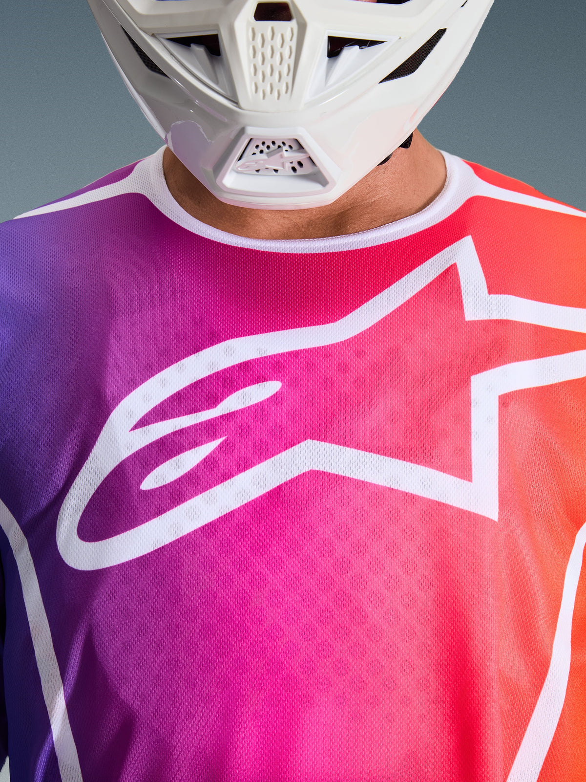 Alpinestars 2026 Fluid Apex Jersey, Motocross-Trikot, Farbverlauf in Violett, Pink und Orange, großes weißes Alpinestars Stern-Logo auf der Brust, Nahaufnahme mit weißem Helm-Kinnschutz