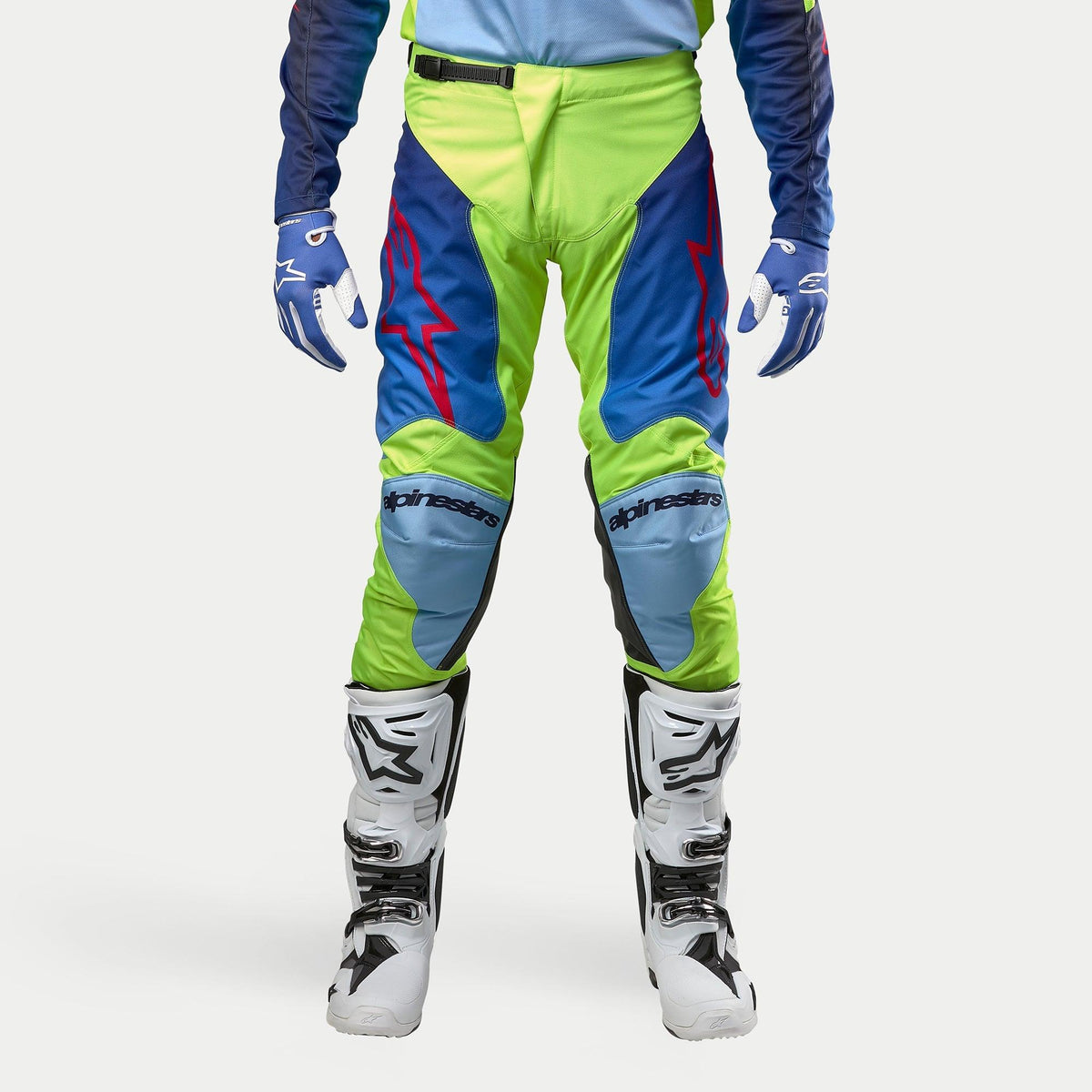 Alpinestars 2024 Racer Hoen broek, motorcrossbroek, fluoriserend geel en nachtblauw, gedragen met witte en zwarte motorcrosslaarzen en blauwe handschoenen, voorzien van opvallende rode logo's, lichtblauwe kniepanelen en een sportief ergonomisch ontwerp voor off-road rijden