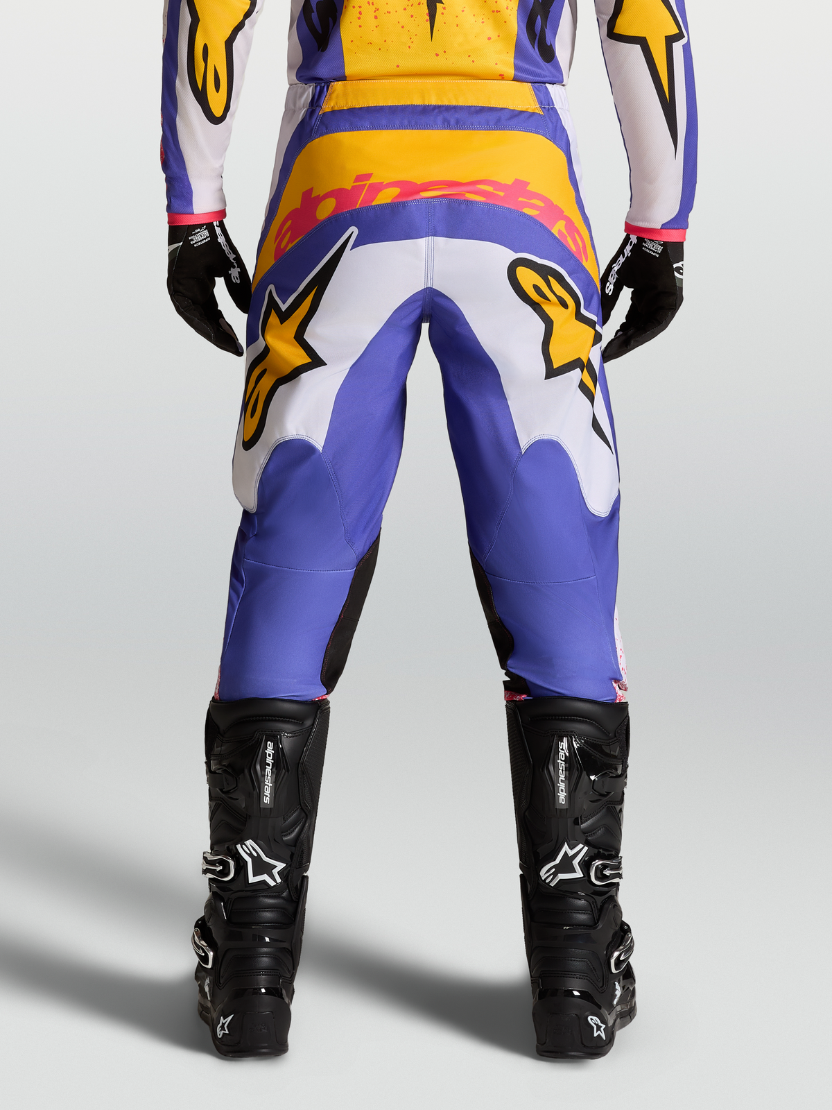 Alpinestars Pantalon Fluid Drag, pantalon de motocross bleu et doré, vue de dos, panneaux en velours côtelé extensible, avec bottes techniques noires.