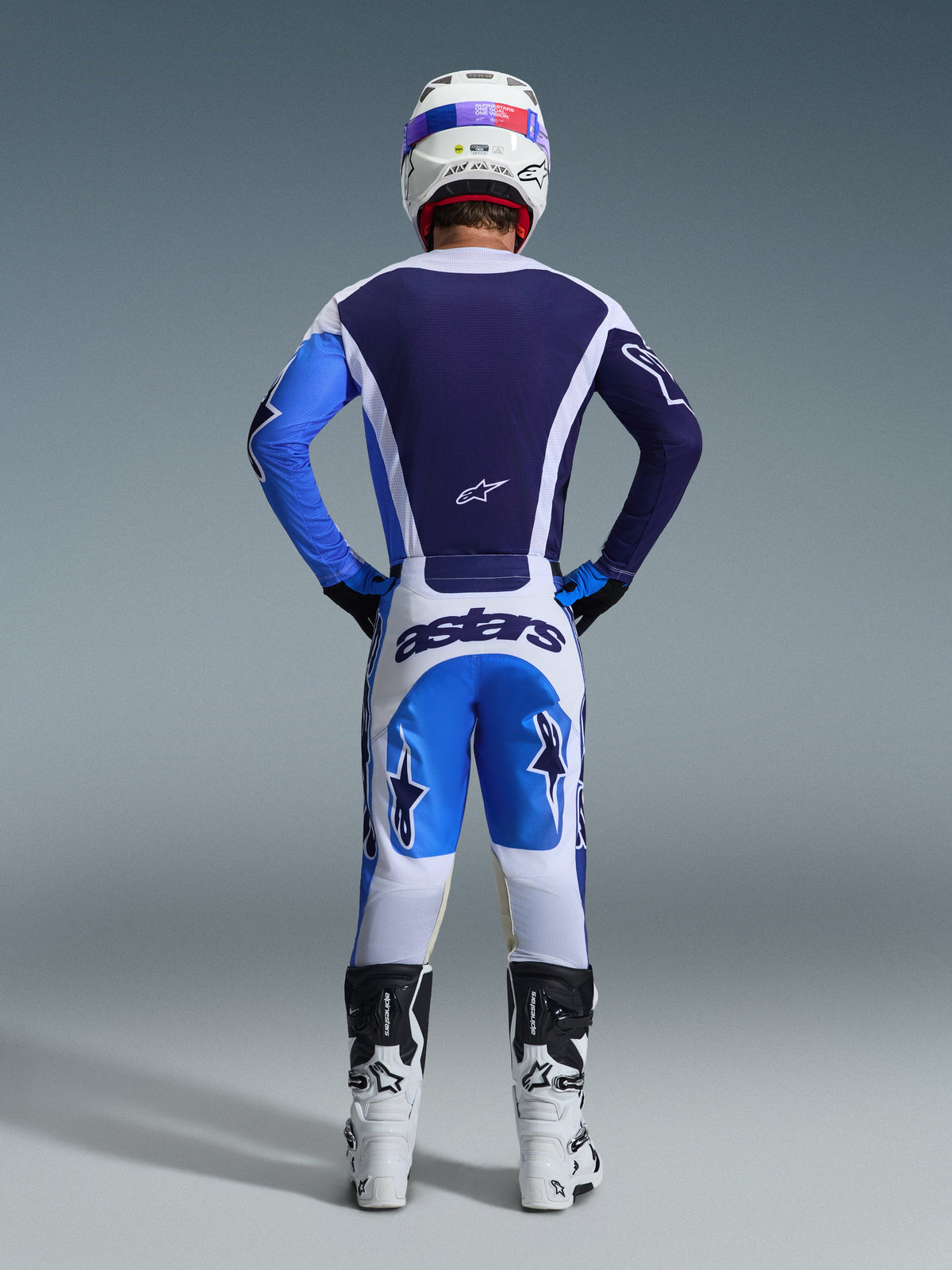 Motocross-Fahrer von hinten in Alpinestars 2026 Racer Air Portl Gear Set, dunkel- und hellblaues Trikot mit weißen Akzenten, weiße Hose mit blauen Einsätzen und dunkelblauem 'astars'-Branding, weiß-schwarze Motocross-Stiefel, weißer Rennhelm, koordiniertes Outfit für Offroad-Performance