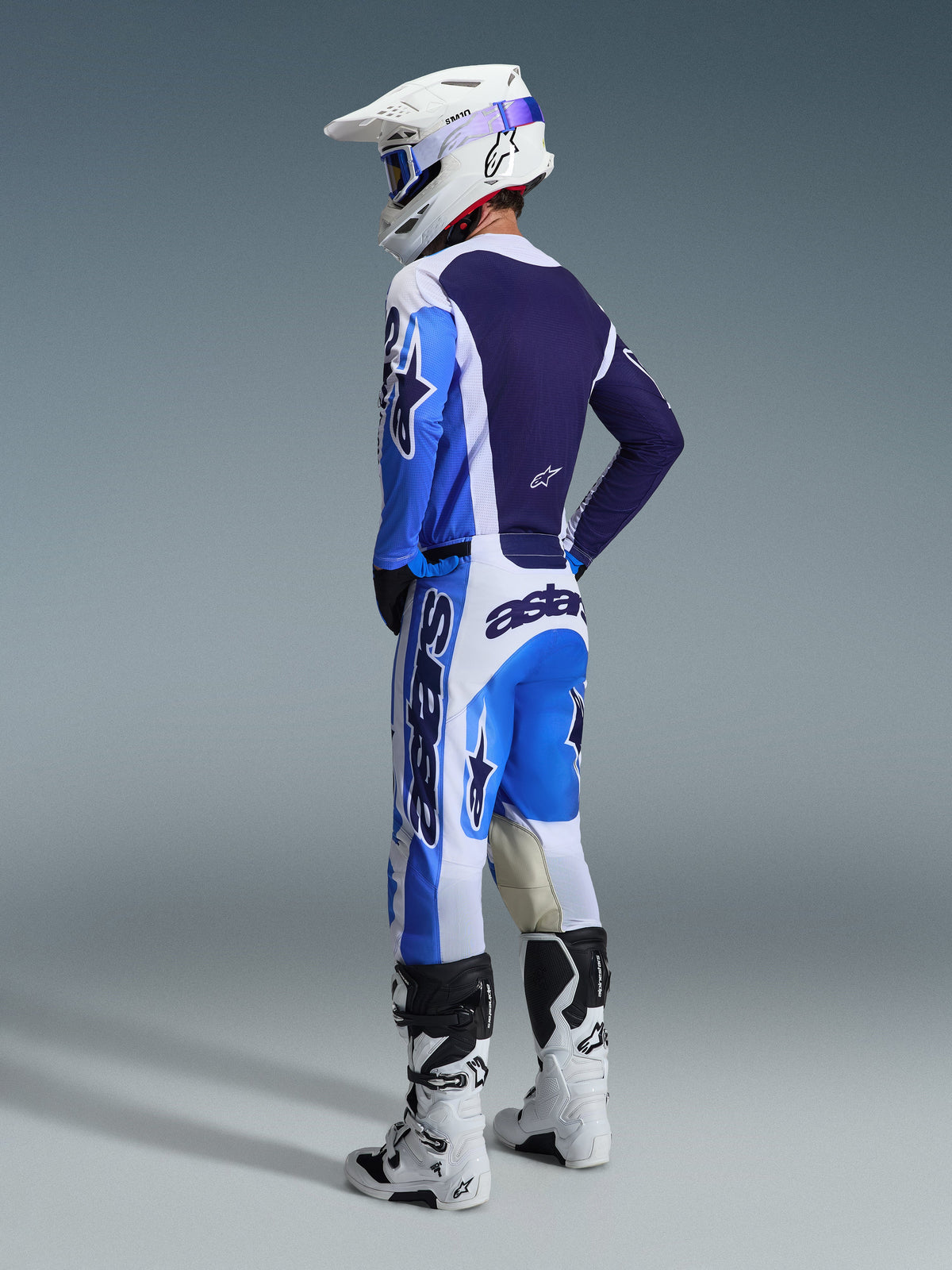 Motocrossrijder van achteren gezien die de Alpinestars 2026 Racer Air Portl Jersey in Ucla Blue en wit draagt, gecombineerd met een bijpassende motocrossbroek, witte Supertech-helm en witte laarzen, wat een volledige gecoördineerde race-uitrusting laat zien.