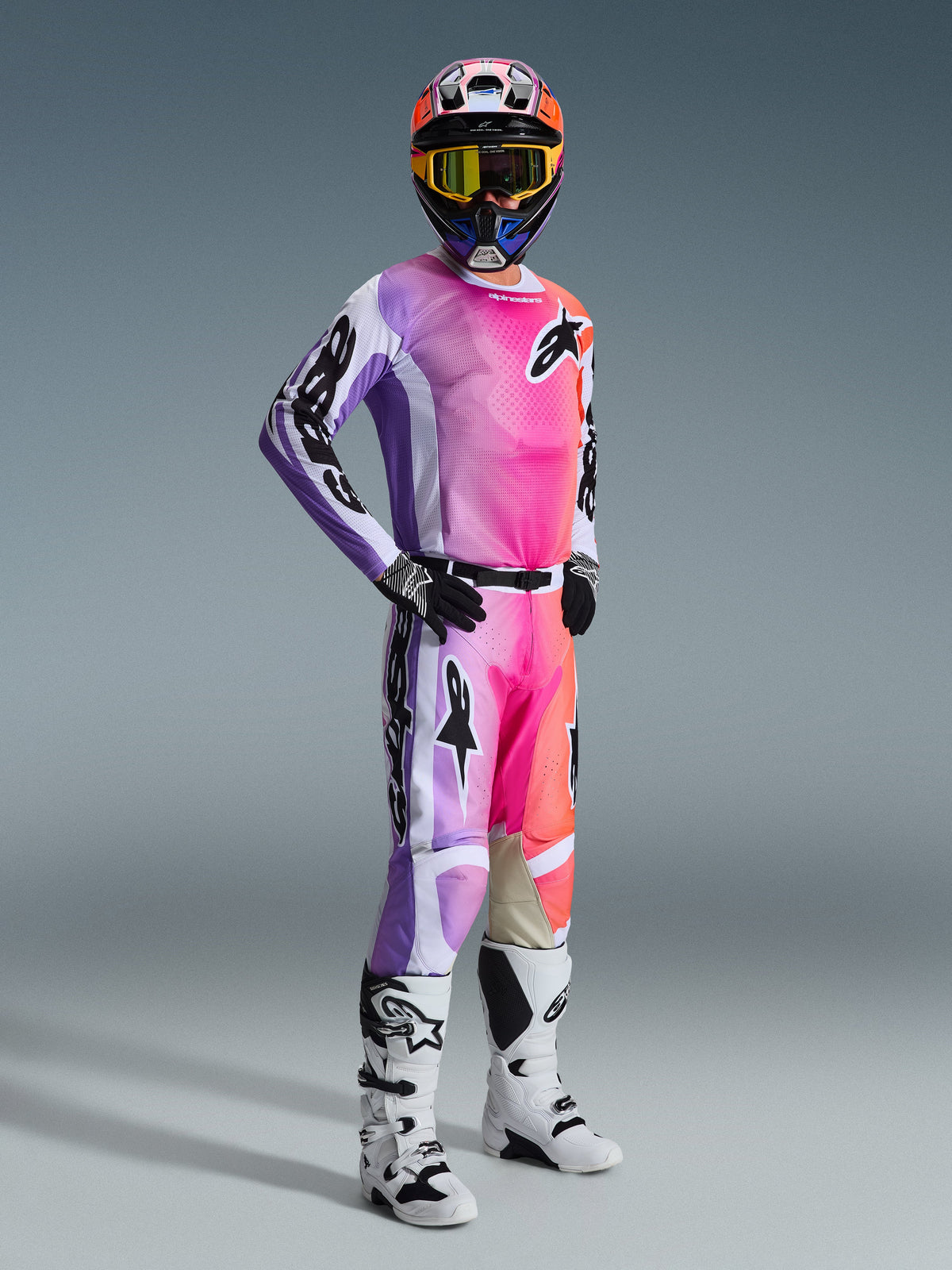 Alpinestars 2026 Racer Air Portl Gear Set, Motocross-Fahrer in voller Rennausrüstung, Jersey und Hose mit leuchtendem Farbverlauf in Lila, Rosa und Orange, schwarze Alpinestars-Logos, weiße und schwarze Akzente, koordinierter mehrfarbiger Helm mit gelb getönter Brille, weiß-schwarze Motocross-Stiefel, schwarz-weiße Handschuhe