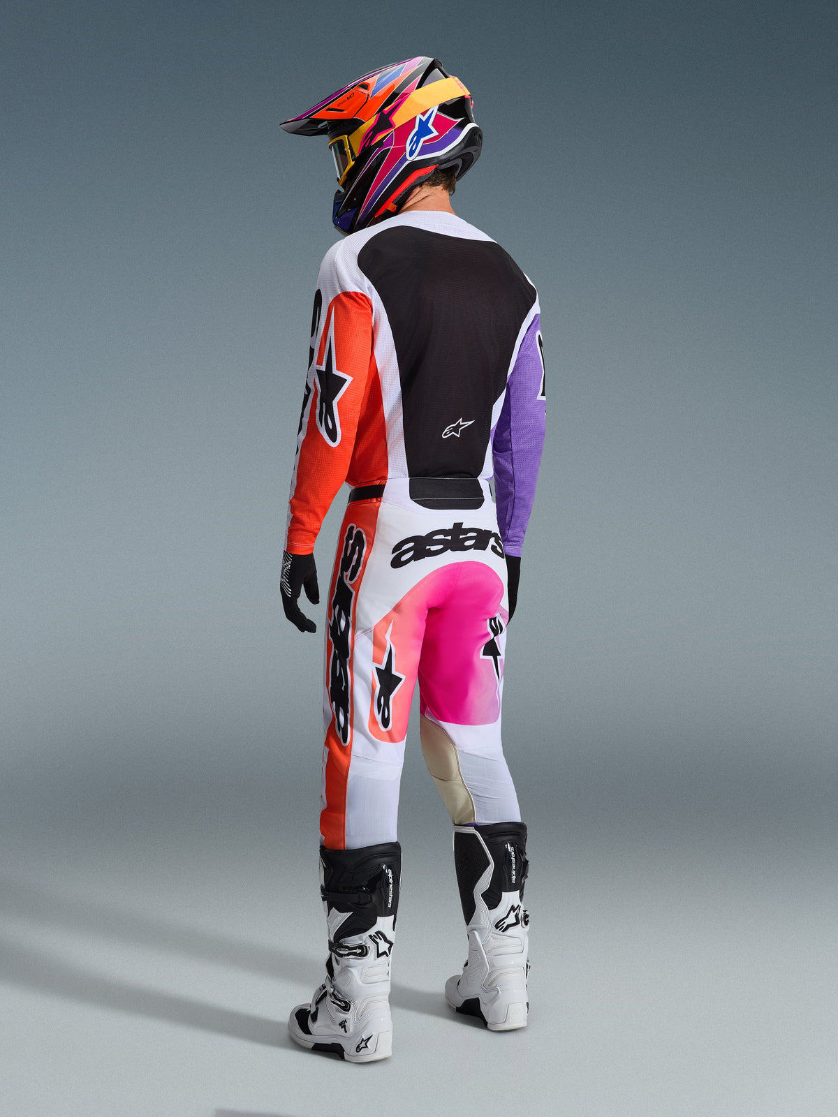 Motocross-Fahrer von hinten im 2026 Racer Air Portl Gear Set, weißes Jersey mit schwarzer Rückenpartie und asymmetrischen orangefarbenen und lila Ärmeln, weiße Hose mit pinkfarbenem Farbverlauf am Gesäß und orangefarbenen Seiteneinsätzen, passender mehrfarbiger Helm und weiß-schwarze Stiefel, professionelle Offroad-Rennausrüstung.