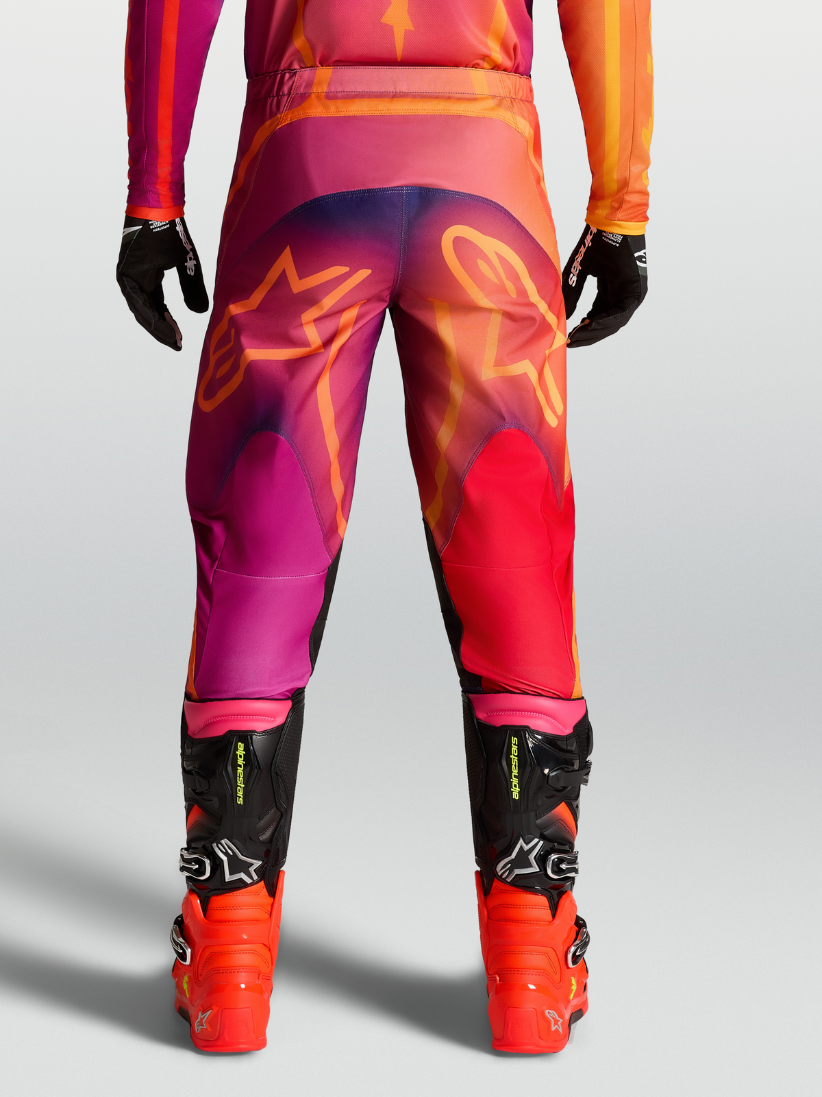 Pantalon Fluid Kepler d'Alpinestars, pantalon de motocross pour homme en violet et orange avec motifs d'étoiles à l'arrière et bottes de racing noires.