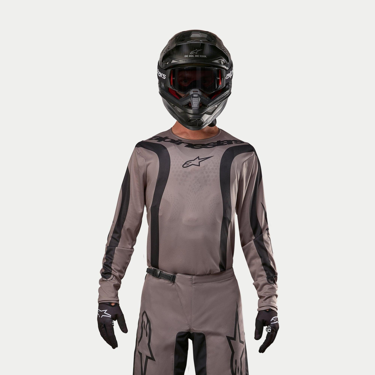 Motorcrosser in de Alpinestars 2024 Fluid Lurv Jersey in Mud Black, een aardse grijsbruine stof met zwarte grafische strepen en een sterlogo op de borst, gecombineerd met een zwarte helm, rode crossbril en zwarte handschoenen voor een gecoördineerde off-road race-look