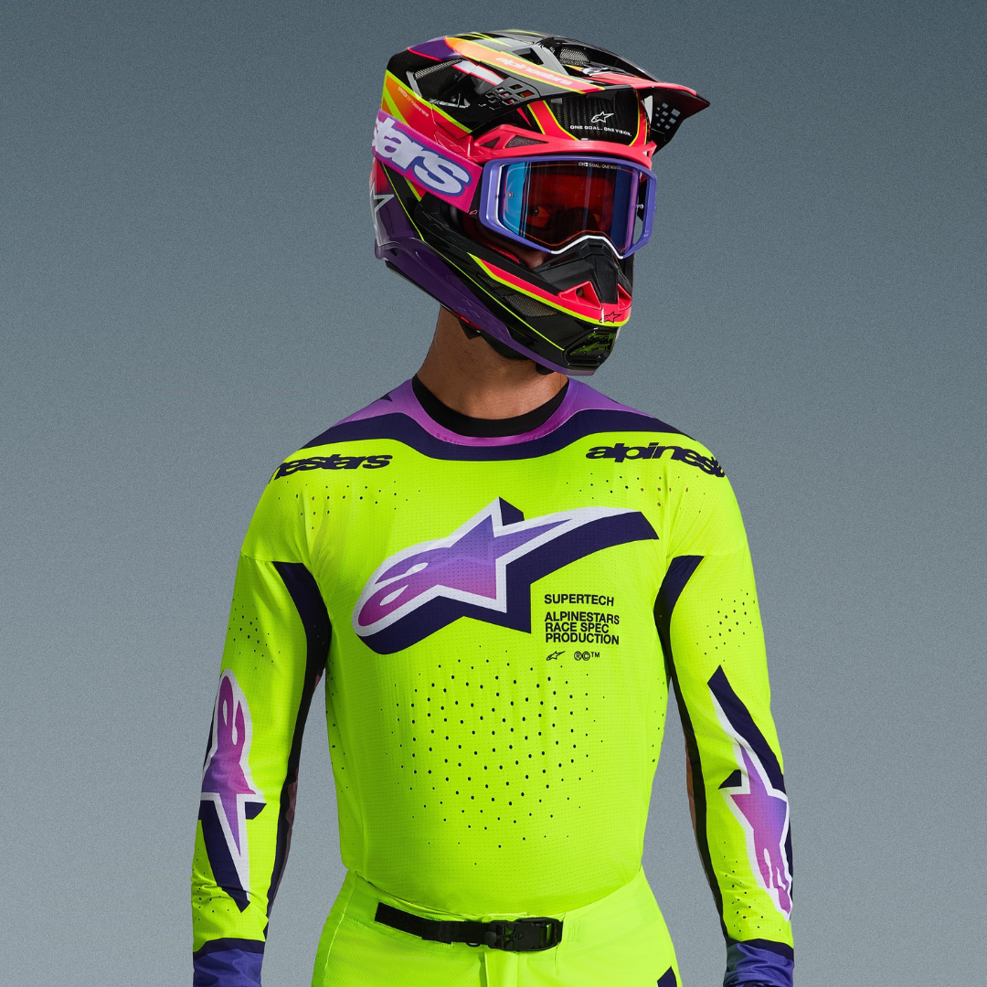 Pilote de motocross portant l'équipement Alpinestars 2026 Supertech Vista, maillot jaune fluo avec détails violets et noirs, grand logo dégradé violet sur la poitrine, casque multicolore éclatant assorti et lunettes violettes, mettant en avant les ventilations découpées au laser et une coupe athlétique professionnelle.