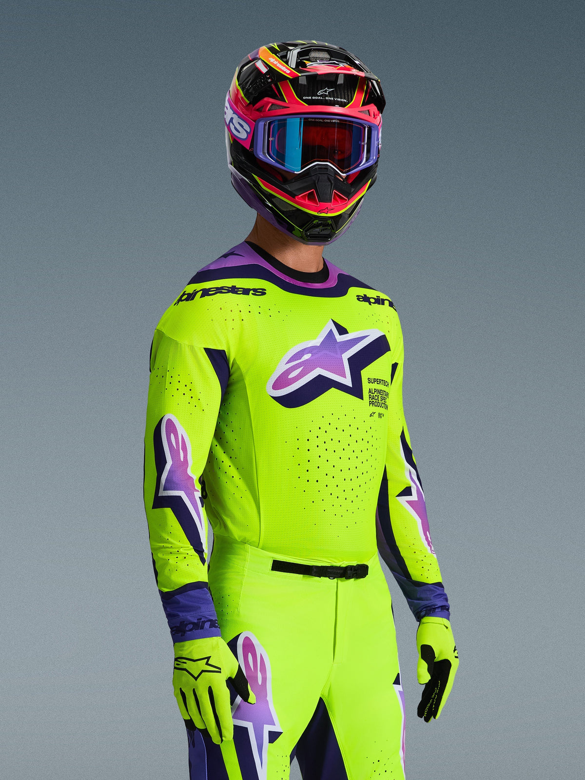 Pilote de motocross portant le maillot Alpinestars 2026 Supertech Vista et la tenue assortie, jaune vif avec accents violets et noirs, arborant un grand logo Astar violet sur la poitrine et les manches, gants assortis et casque de course multicolore avec lunettes, coupe athlétique de niveau pro pour la compétition tout-terrain