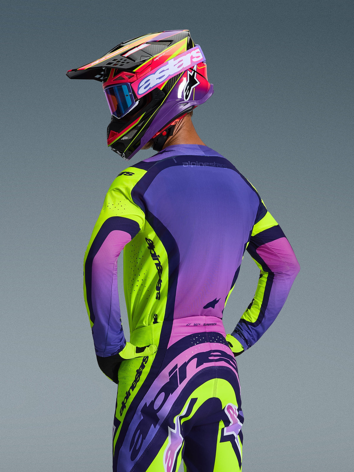 Pilote de motocross vu de dos portant le maillot Alpinestars Supertech Vista 2026 et le pantalon assorti, coloris Jaune Violet Noir avec dégradés vibrants, casque de course coordonné avec lunettes bleues et gants jaunes, tenue de course tout-terrain professionnelle avec coupe athlétique et logos intégrés.