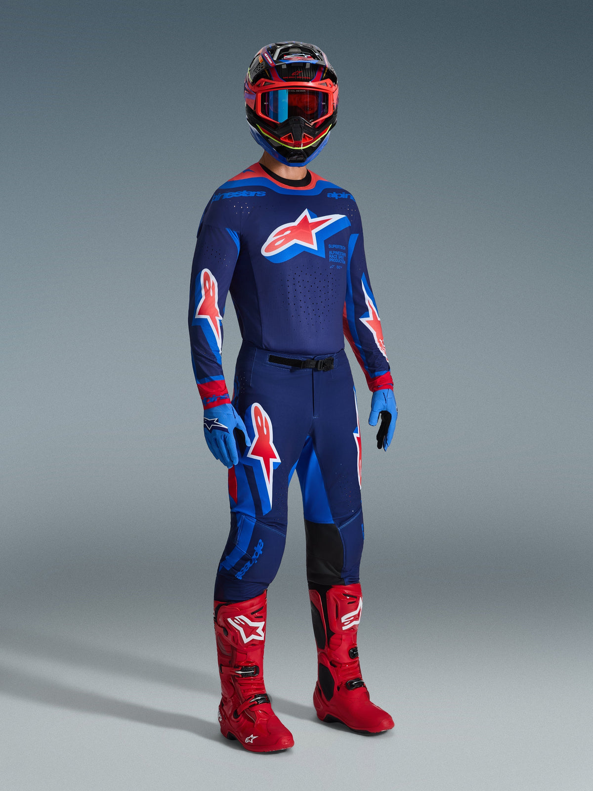 Motorcrosser in volledige race-uitrusting, met de Alpinestars 2026 Supertech Vista Jersey in Dark Navy, Hot Coral en Cobalt, bijpassende crossbroek met opvallende ster-logo's, felrode motorcrosslaarzen, blauwe handschoenen en een veelkleurige helm met rode bril, professionele off-road racetenue.
