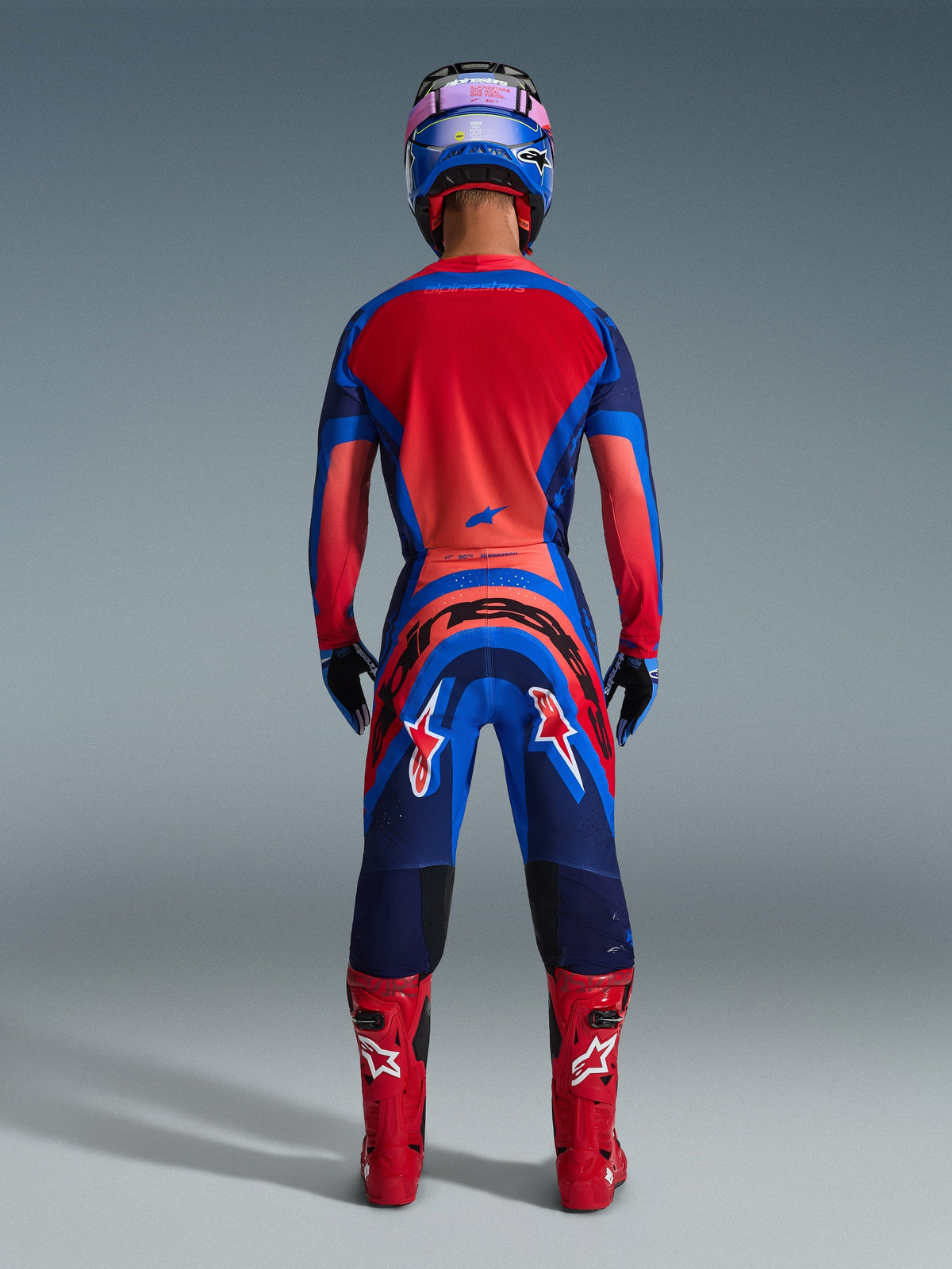 Motocrossrijder van achteren gezien in de Alpinestars 2026 Supertech Vista Jersey in Dark Navy, Hot Coral en Cobalt, bijpassende kledingset met broek, rode racelaarzen en een blauwe helm, professionele off-road racekleding.