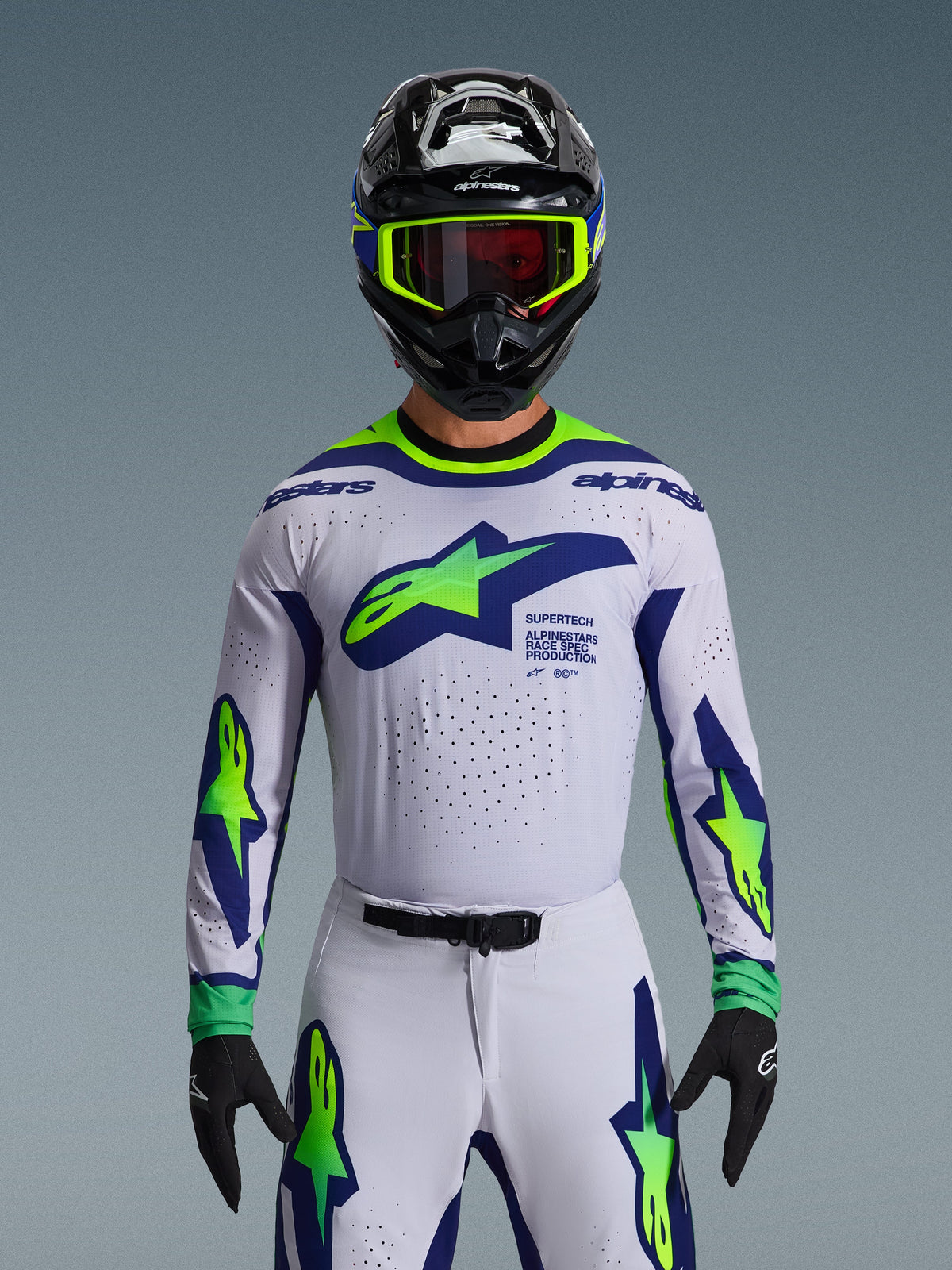 Ensemble d'équipement Alpinestars Supertech Vista 2026, maillot et pantalon de motocross, blanc avec graphismes bleus et vert fluo, porté avec un casque noir et des lunettes fluo, avec ventilation découpée au laser et coupe athlétique pour la compétition professionnelle.