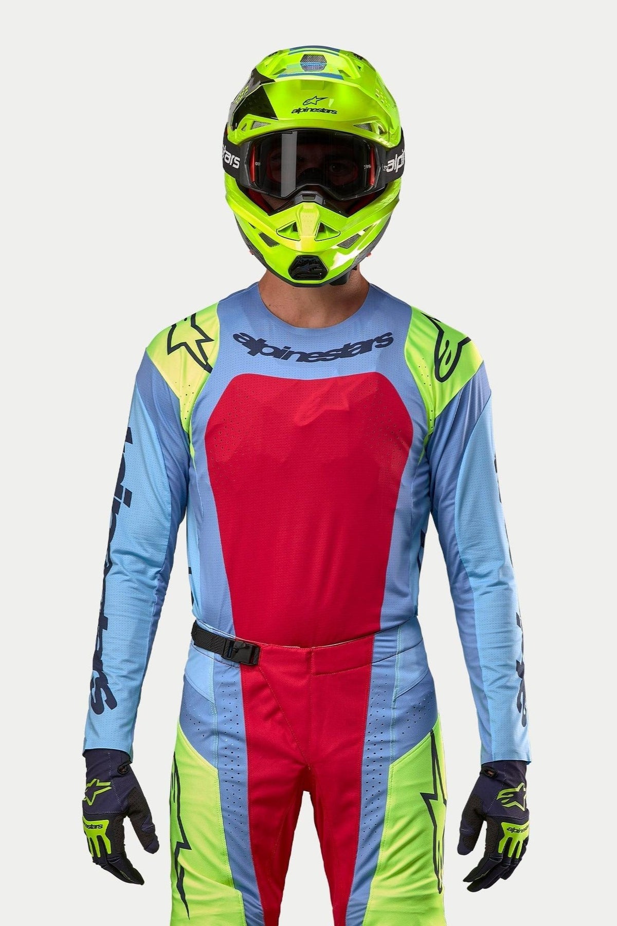 Pilote de motocross portant une tenue de course complète, maillot Techstar Ocuri 2024 bleu clair avec un large panneau de poitrine rouge et des accents jaune fluo aux épaules, casque jaune fluo assorti, pantalon coordonné rouge et jaune fluo, tenue professionnelle pour la course tout-terrain