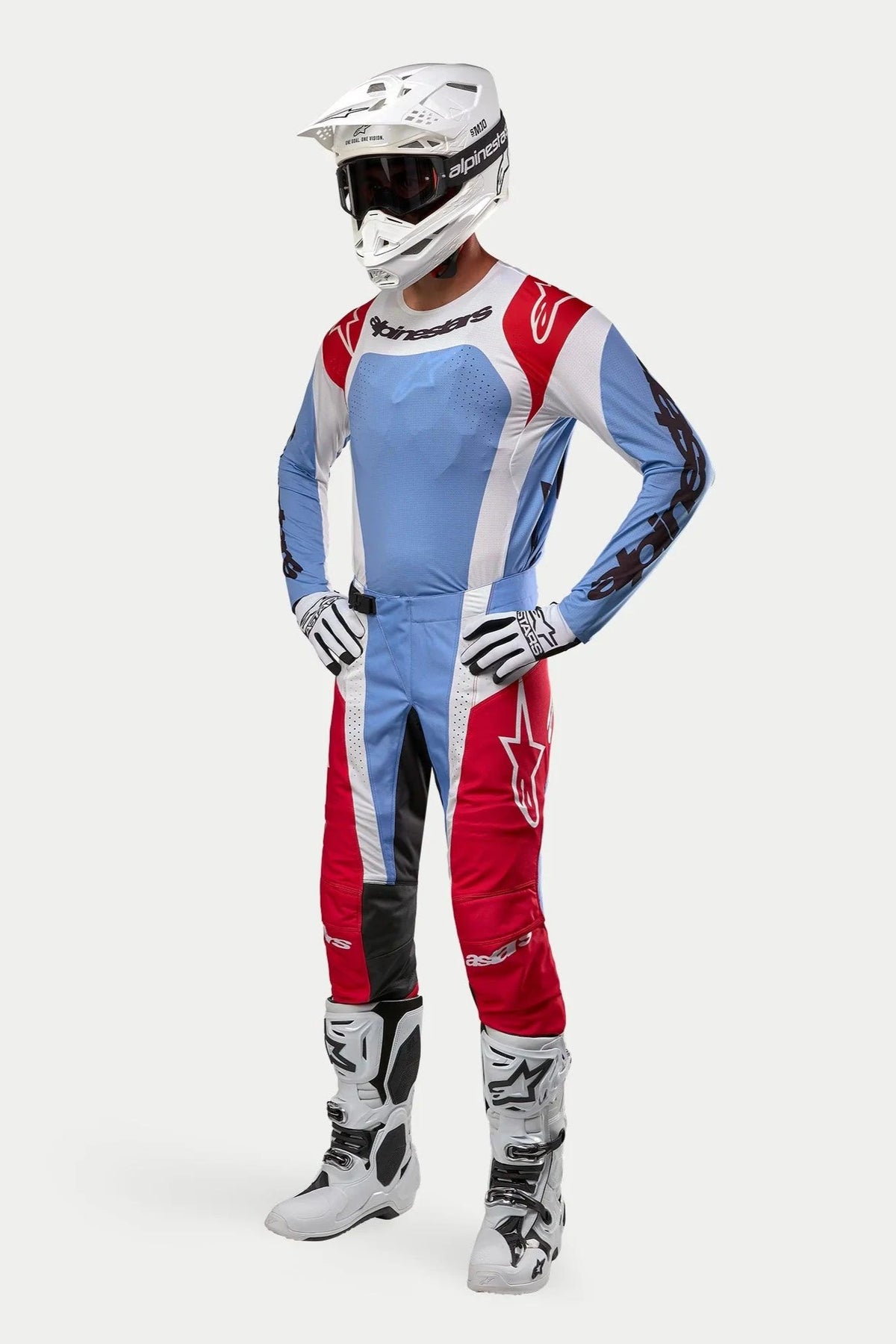 Pilote de motocross portant l'équipement Alpinestars Techstar Ocuri 2024, maillot et pantalon bleu clair, rouge Mars et blanc, casque blanc avec lunettes, gants et bottes blancs et noirs, tenue de course professionnelle pour performance tout-terrain