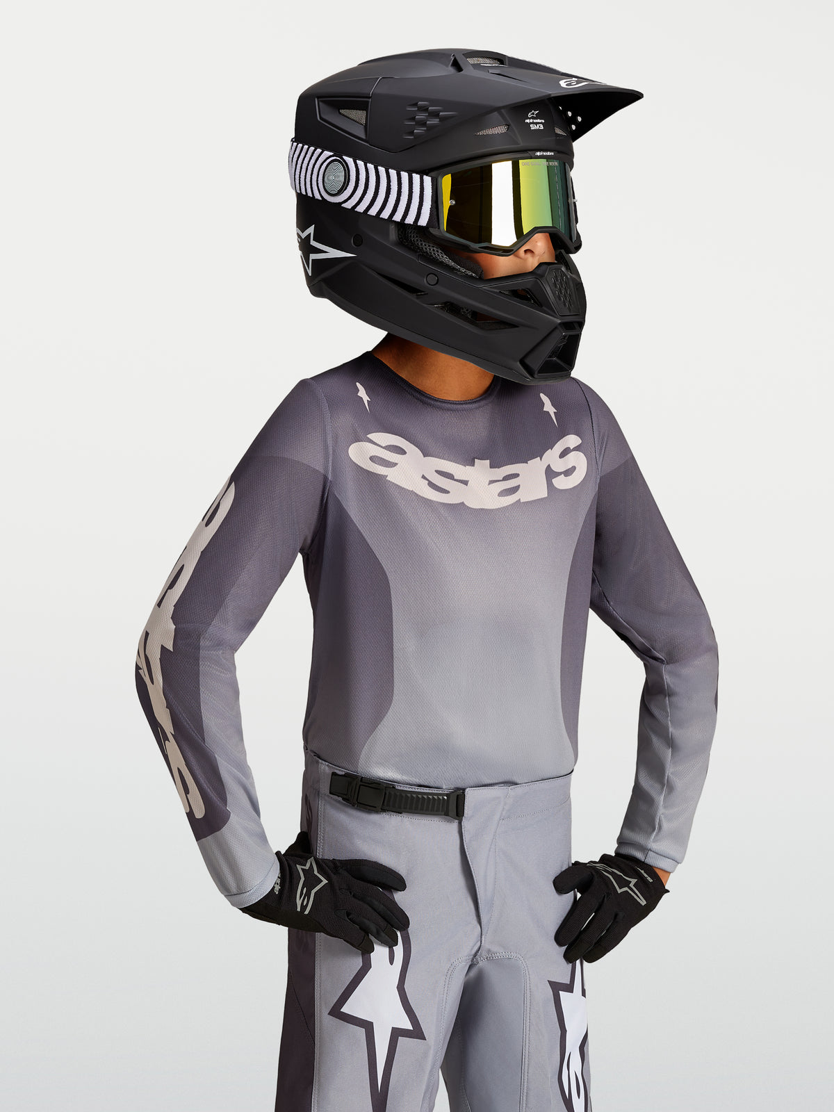 Alpinestars Fluid Jugend-Trikot Amber 2027, Jugend-Motocross-Trikot, Grau und Sand, leichtes atmungsaktives Material, getragen mit schwarzem Rennhelm und Handschuhen.