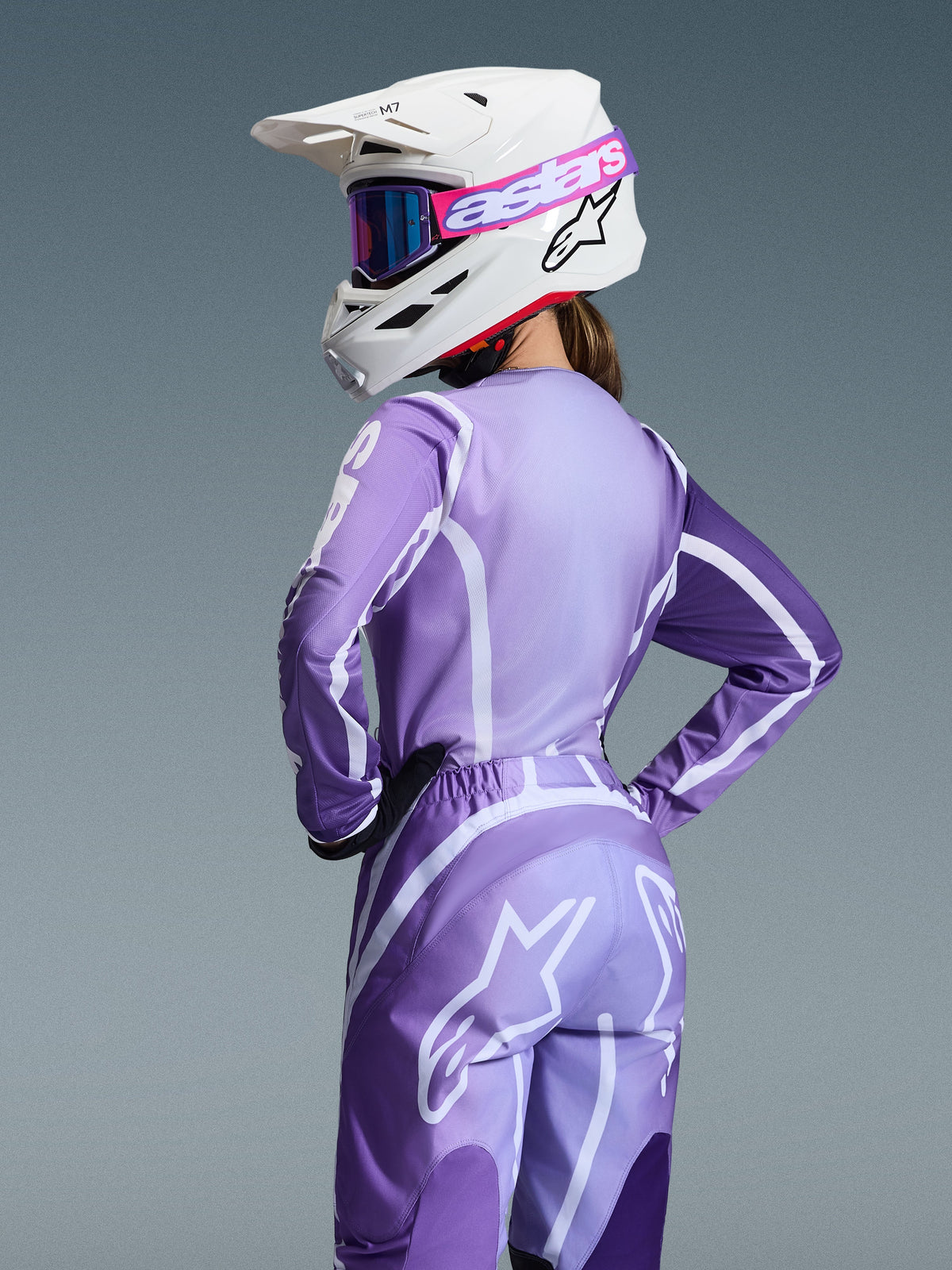 Motorcrossrijder in Alpinestars 2026 Stella Fluid Apex Gear Set, paarse en lavendelkleurige jersey en broek met witte graphics, witte helm met roze en paarse crossbril, specifieke damespasvorm, gecoördineerde uitrusting voor off-road racen