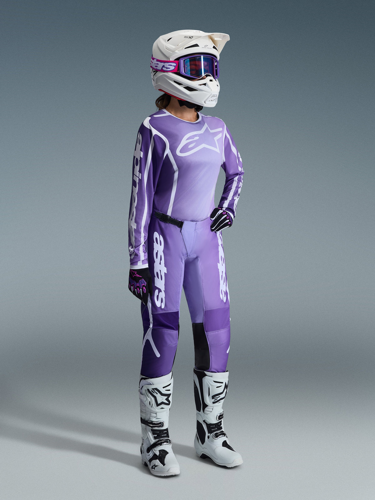 Alpinestars 2026 Stella Fluid Apex Gear Set, paars en lavendel, vrouwelijke motorcrossrijder met bijpassende jersey en broek, witte helm met paarse crossbril, witte en zwarte laarzen, zwarte en paarse handschoenen, complete performance-outfit voor dames off-road racing