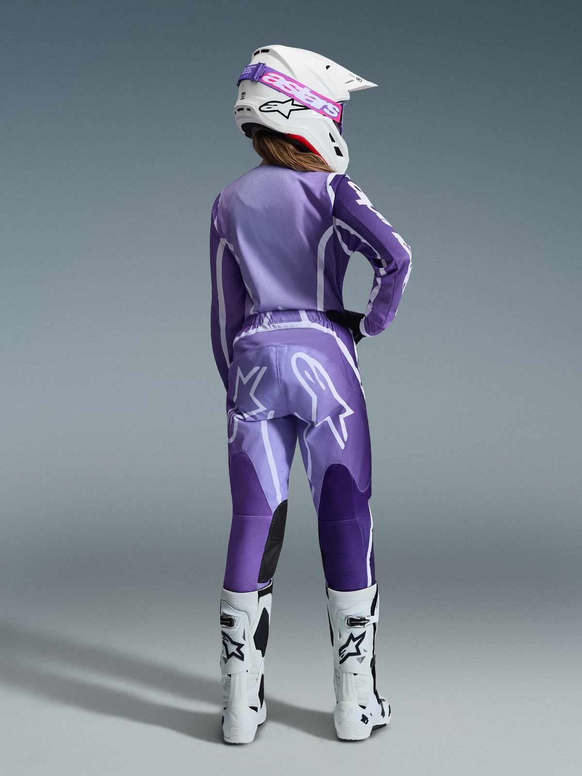Alpinestars 2026 Stella Fluid Apex Gear Set, motocrosskleding voor dames, paars en lavendel met witte accenten, vrouwelijke rijder van achteren gezien in bijpassende jersey en broek, witte helm met roze crossbril en witte laarzen, gecoördineerde sportieve race-outfit