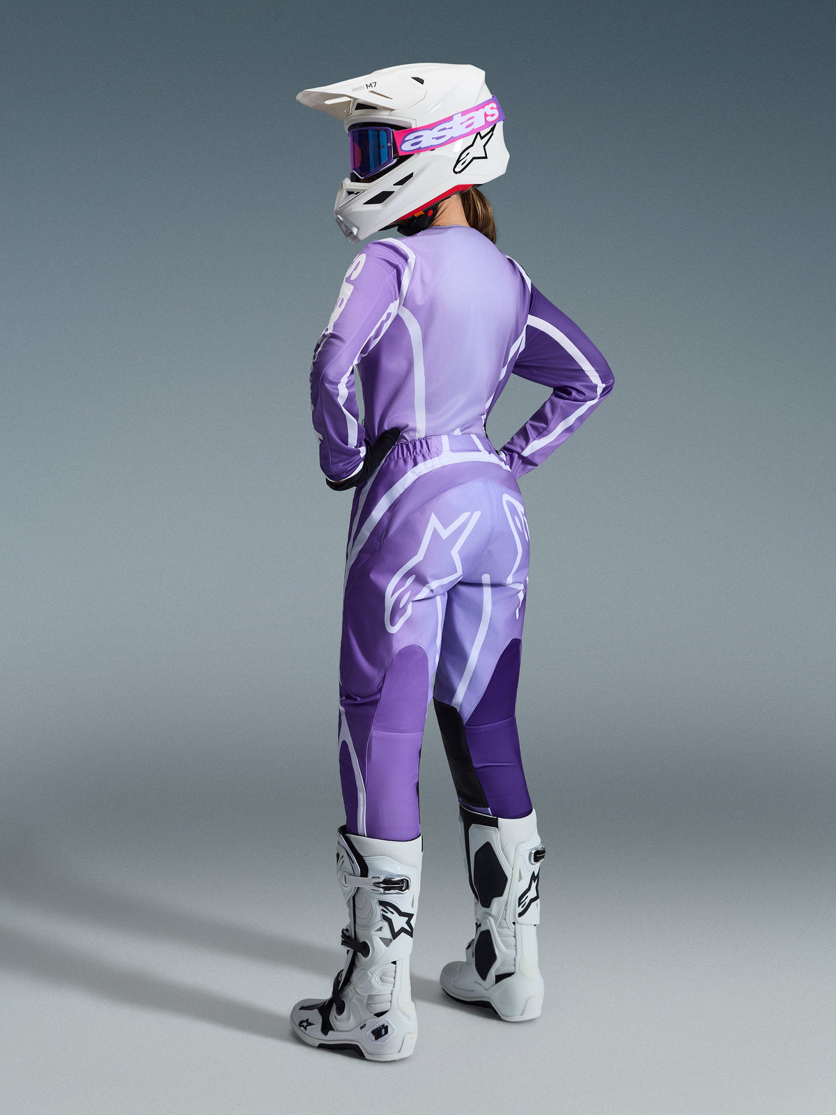 Alpinestars 2026 Stella Fluid Apex Gear Set voor dames, vrouwelijke motorcrosser in paarse en lavendelkleurige jersey en broek met witte accenten, witte helm met roze bril, witte en zwarte laarzen, gecoördineerde outfit voor off-road racing