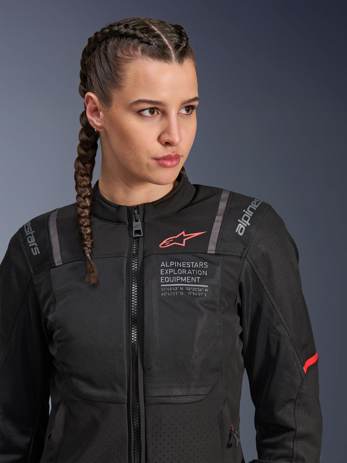 Alpinestars Stella ST-2 Air Jacke, Motorradjacke für Damen in Schwarz, mit Mesh-Einsätzen für Luftzirkulation und „Alpinestars Exploration Equipment“-Schriftzug mit Koordinaten, rotes Logo-Detail auf der Brust, getragen von einem Model mit geflochtenem Haar.