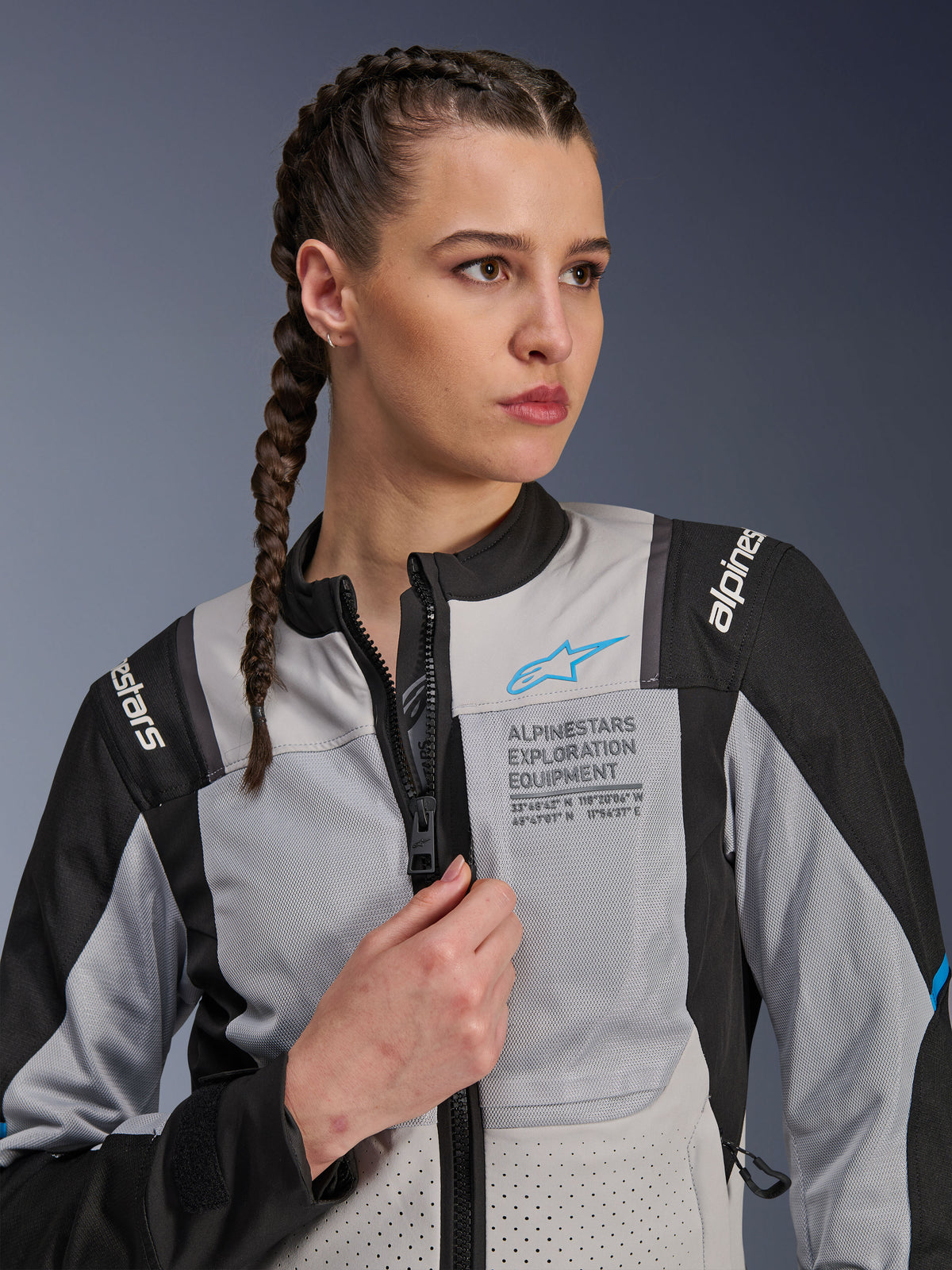 Vrouw die de Alpinestars Stella ST-2 Air Jacket in Ice Gray en Black draagt, close-up portret, met mesh-panelen voor ventilatie, 'ALPINESTARS EXPLORATION EQUIPMENT' branding op de borst met een blauw logo, moderne adventure motorjas