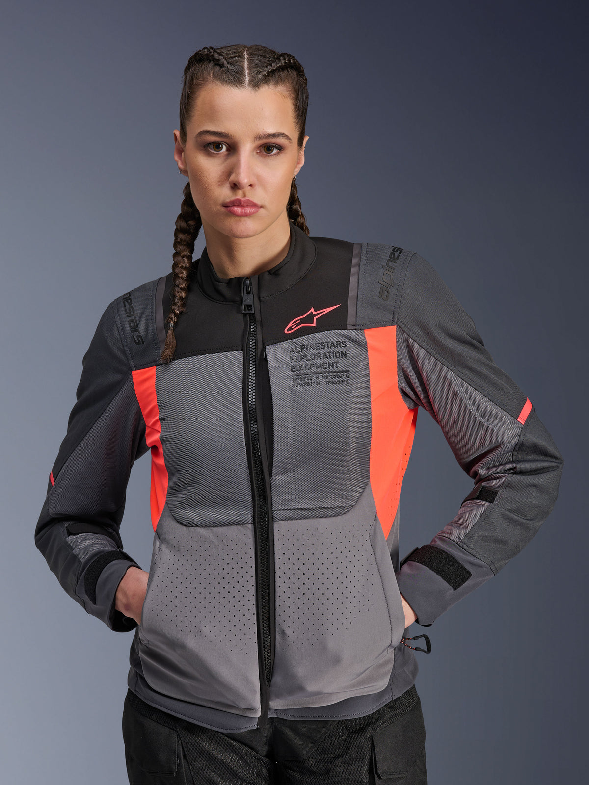 Alpinestars Stella ST-2 Air jas, dames motorjas, donkergrijs en fluo koraal, gedragen door model met ingevlochten haar, voorzien van technische mesh-panelen voor luchtstroom, laserperforaties en 'Alpinestars Exploration Equipment'-branding op de borst, zomerse toeruitrusting