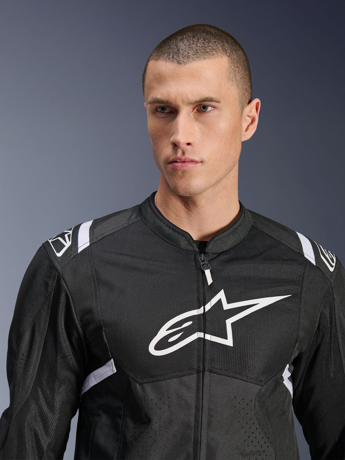 Alpinestars T-SPS Air V2 Jas, textiel motorjas, zwart en wit, voorzien van een groot wit logo op de borst en mesh-panelen, sportieve pasvorm gedragen door een mannelijk model