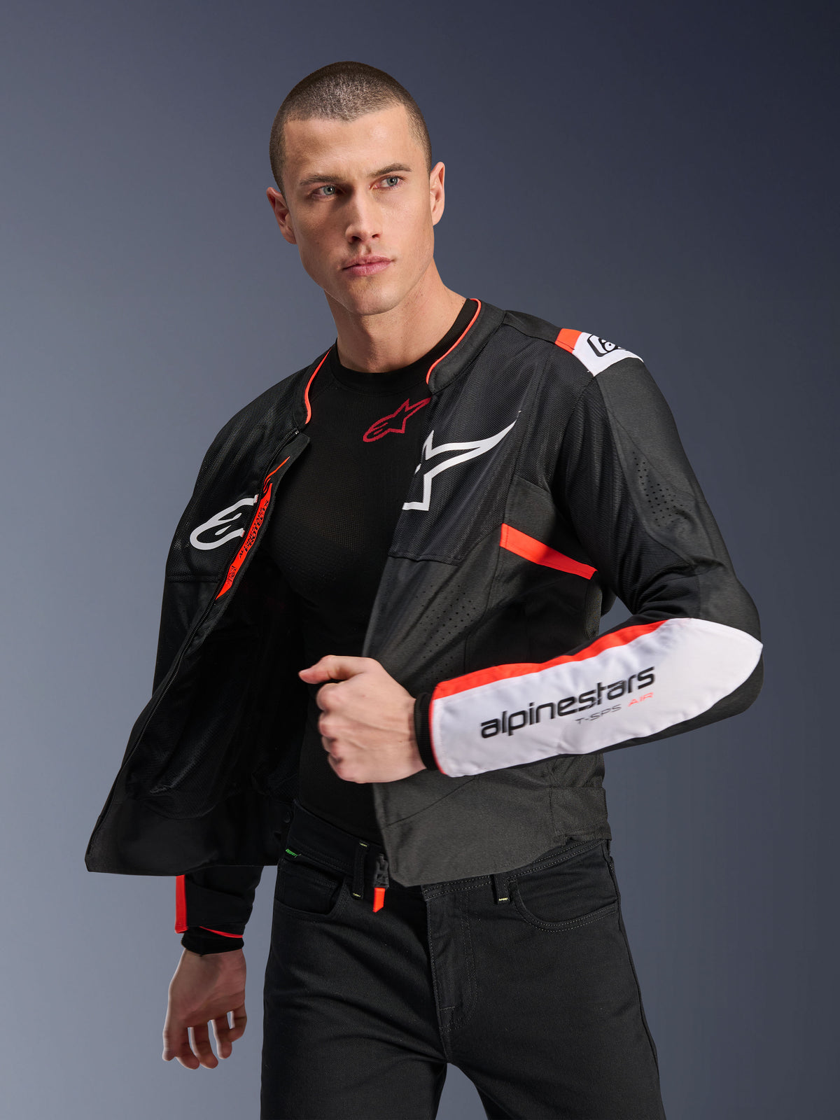 Modèle masculin portant la veste Alpinestars T-SPS Air V2, veste de moto en textile noir avec accents blancs et rouge fluo, dotée de panneaux en mesh pour la circulation de l'air, logo Alpinestars blanc sur la poitrine et marquage sur la manche, portée ouverte sur une couche de base noire