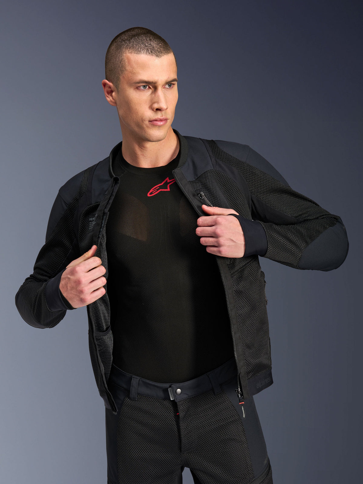 Alpinestars Troop-Air Pro Jacke, Schwarz, hochwertige technische Air-Jacke mit Protektoren und Mesh-Konstruktion, offen getragen über einem schwarzen Base-Layer mit rotem Logo, kombiniert mit passender Mesh-Motorradhose, urbaner zeitgenössischer Fahrstil