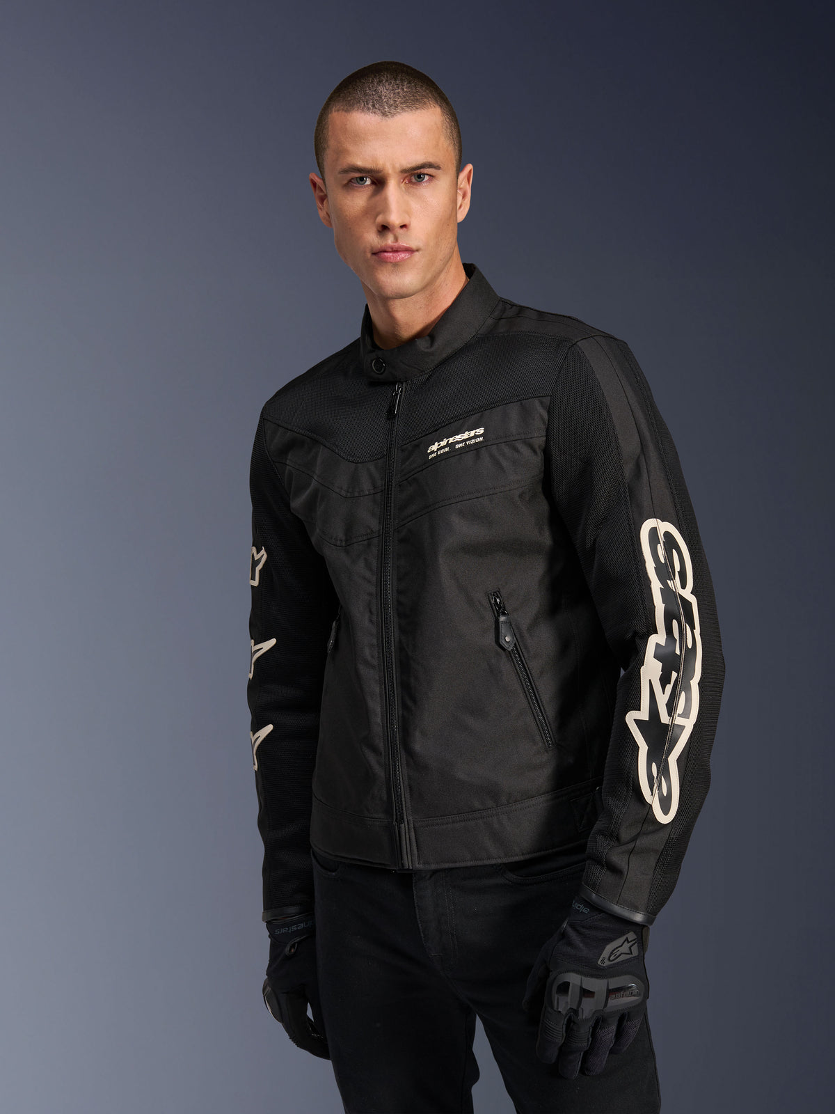 Alpinestars T-Dyno Air Jacke in Schwarz, männliches Model trägt eine Vintage-inspirierte Textil-Motorradjacke mit Mesh-Einsätzen zur Belüftung, großes weißes Alpinestars-Logo auf den Ärmeln, kombiniert mit schwarzen Motorradhandschuhen, urbanes Design