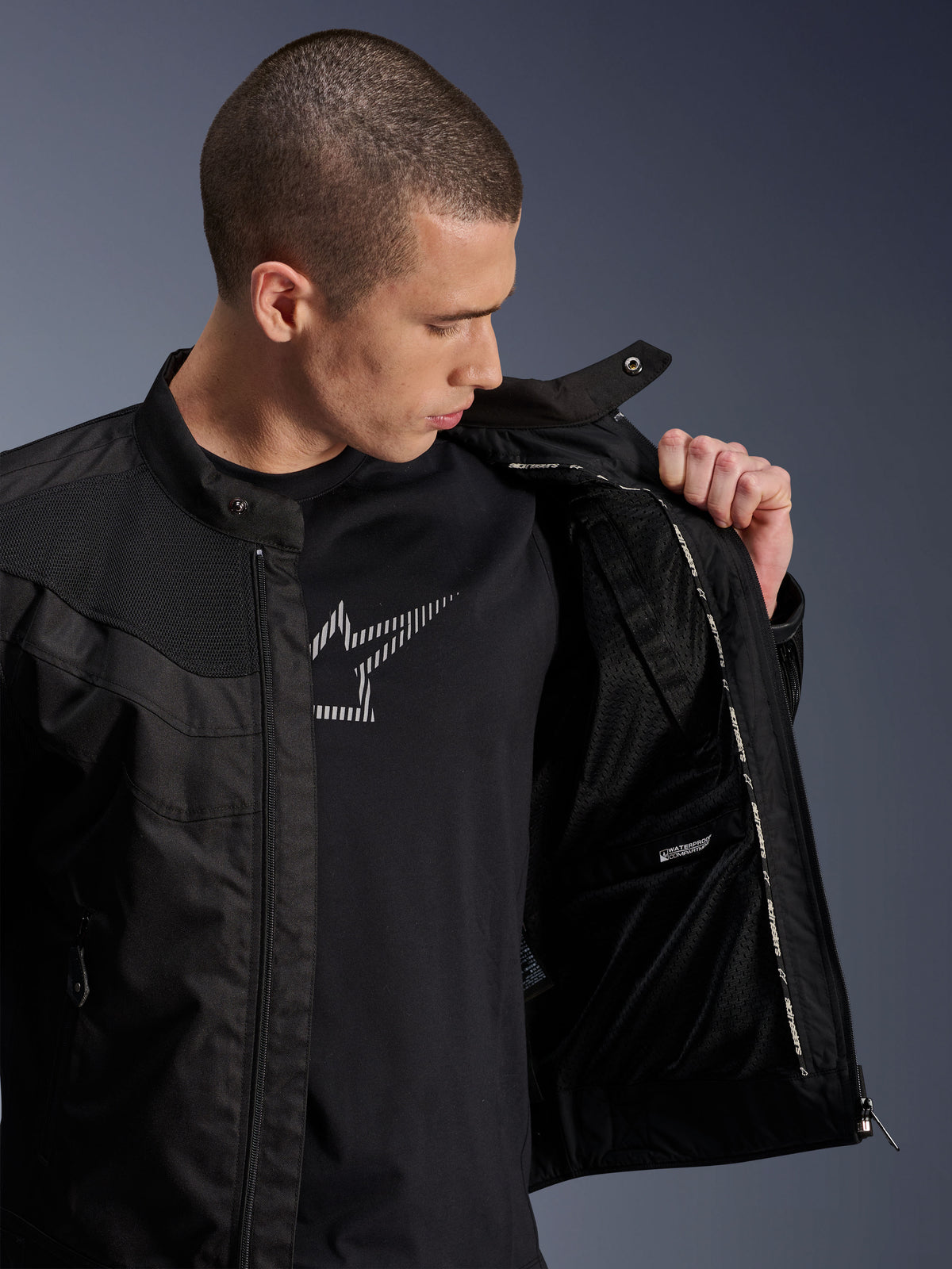 Alpinestars T-Dyno Air Jacke, Schwarz, männliches Model zeigt das Mesh-Innenfutter und das Etikett des wasserdichten Fachs, getragen über einem schwarzen Grafik-T-Shirt, moderne urbane Motorradausrüstung