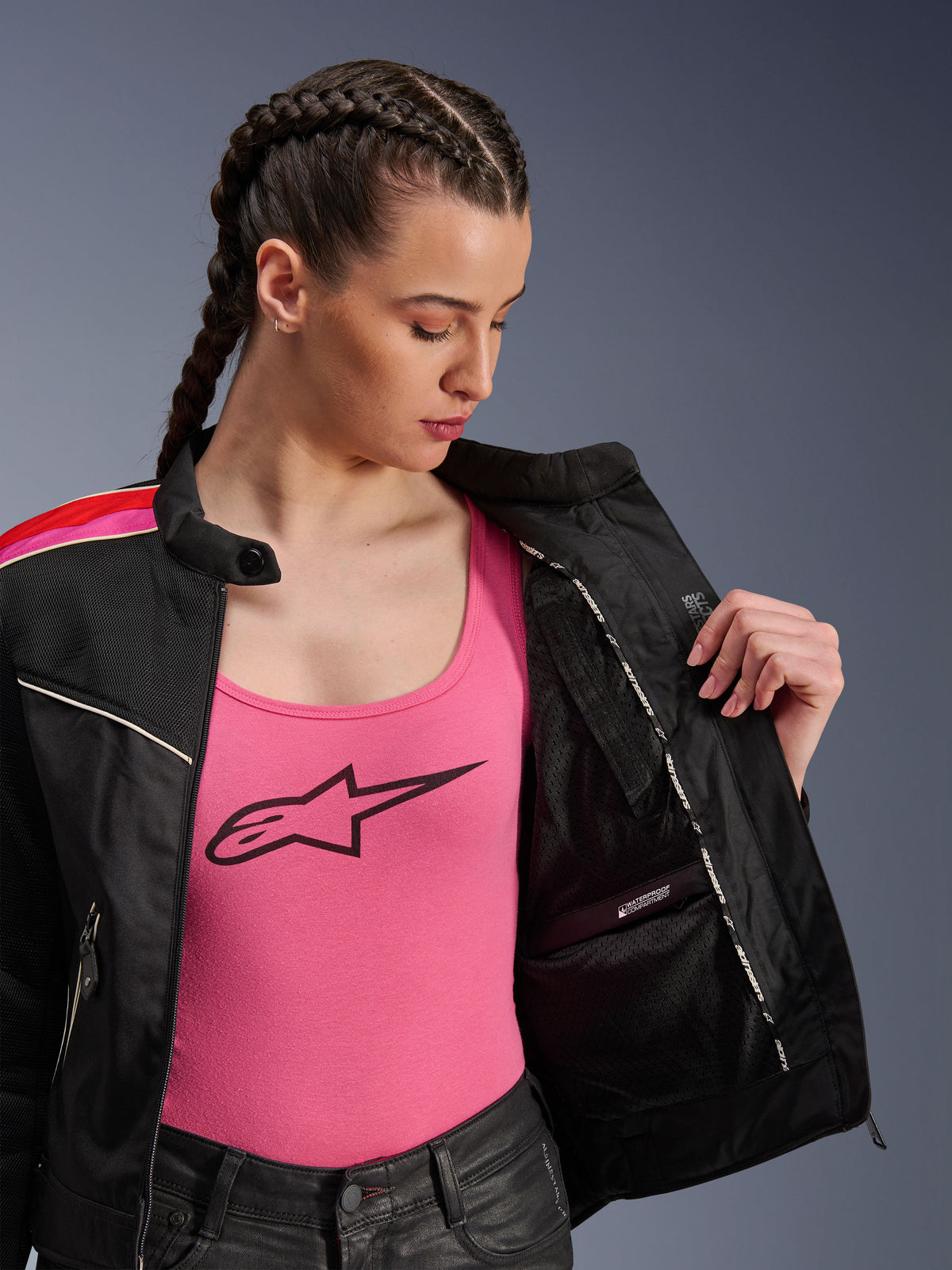 Motorradfahrerin in der Alpinestars Stella T-Dyno Air Jacke in Schwarz, Hellrot und Fuchsia, teilweise geöffnet, um das interne wasserdichte Fach zu zeigen, getragen über einem pinkfarbenen Logo-Tanktop, betont die atmungsaktiven Mesh-Einsätze und die taillierte Biker-Passform.