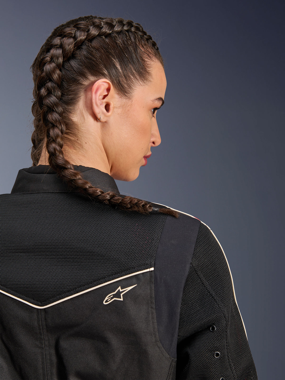 Frau in der Alpinestars Stella T-Dyno Air Jacke, Nahaufnahme der Rückansicht mit Fokus auf Schultern und Passe, Schwarz mit atmungsaktiven 3D-Mesh-Einsätzen, weißer Paspelierung und kleinem Astars-Logo, Modell mit geflochtenem Haar, urbane Motorradbekleidung