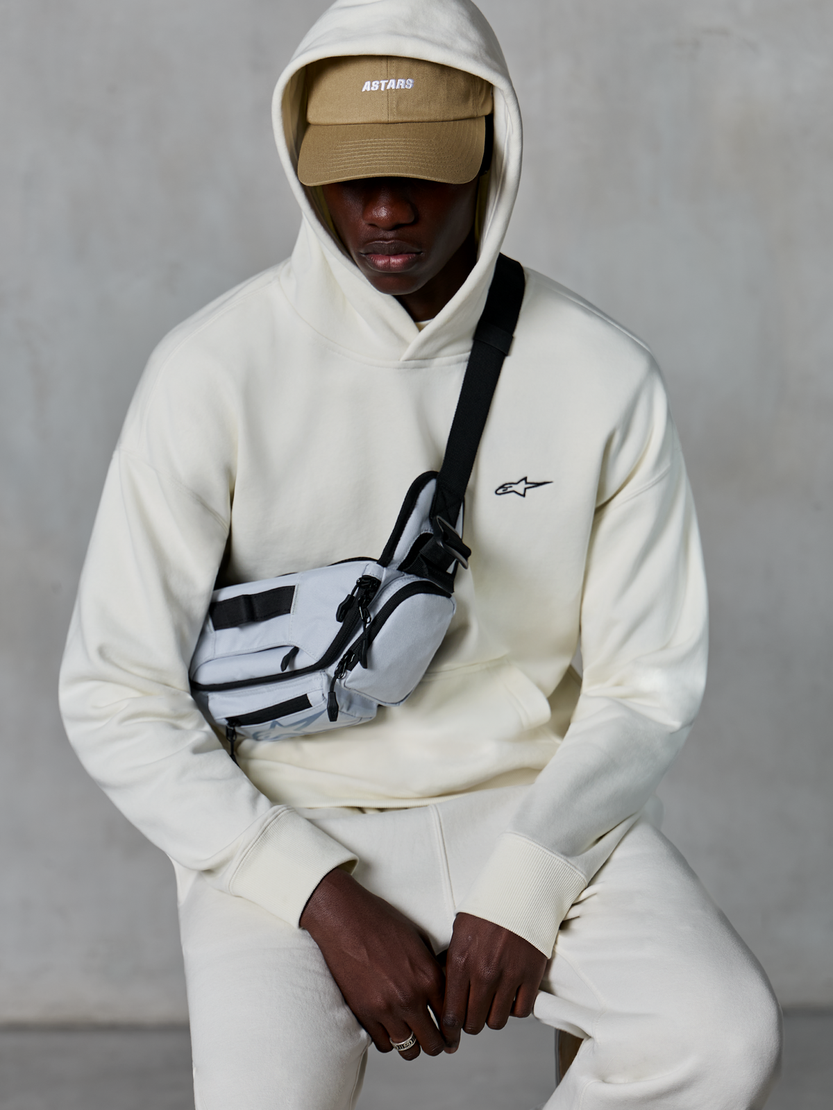 Alpinestars Essentials Ageless Hoodie, casual streetwear, Off White, klein zwart geborduurd logo op de borst, capuchon gedragen over een beige pet met ASTARS-branding, gecombineerd met een lichtgrijze cross-body tas, premium lifestyle kleding
