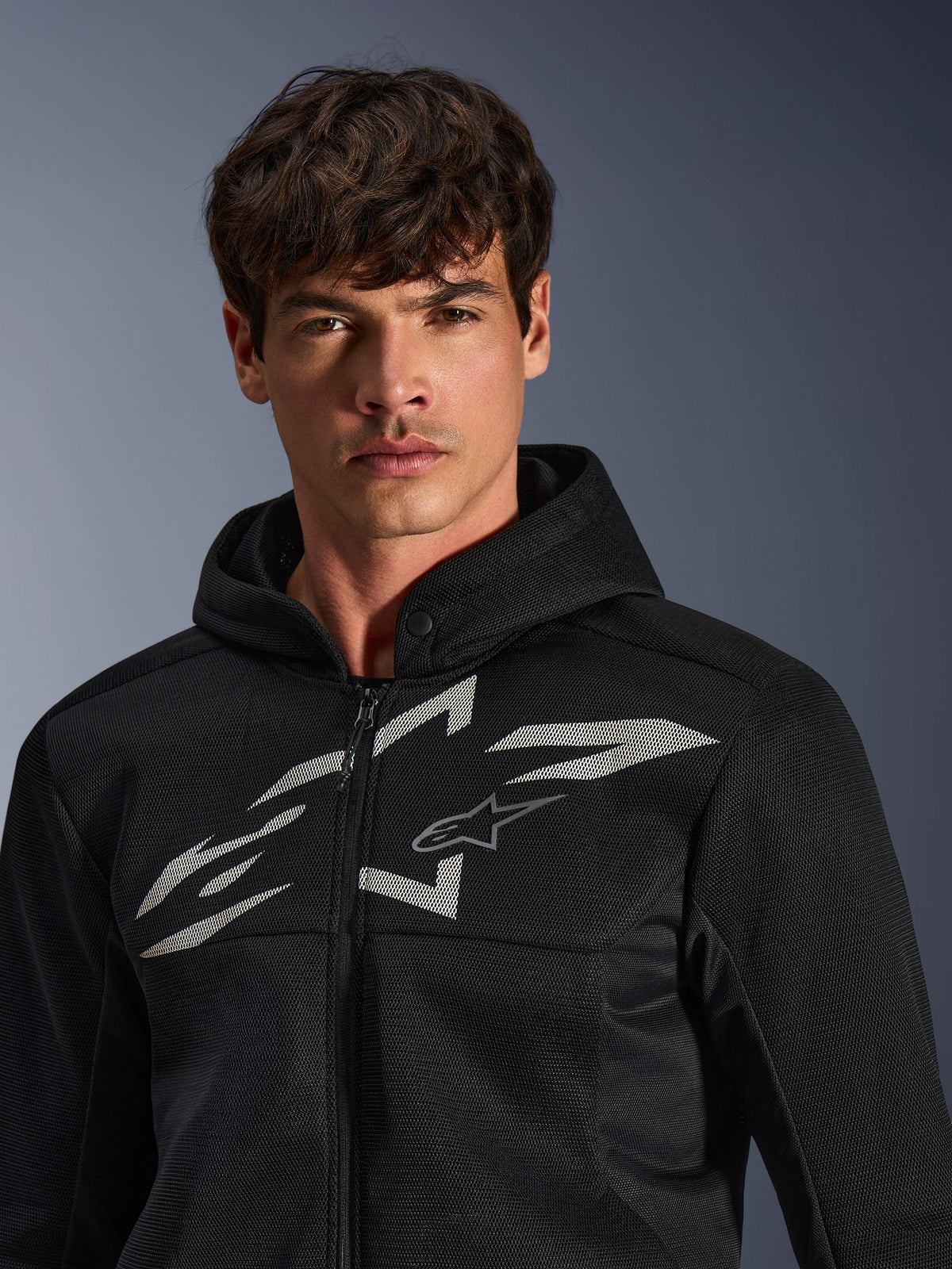 Alpinestars Chrome Superairflow Hoodie, schwarzer Motorrad-Hoodie, verfügt über eine 3D-Mesh-Konstruktion mit einem großen weißen Grafik-Logo auf der Brust, von einem Model getragen, urbane Sport-Fahrbekleidung mit modernem, belüftetem Design