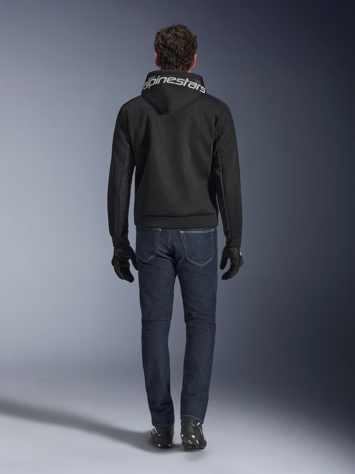 Alpinestars Chrome Superairflow Hoodie, schwarzer Motorrad-Hoodie mit weißem Branding auf der Kapuze, Rückansicht, getragen mit dunkelblauen Jeans, schwarzen Handschuhen und schwarz-weißen Stiefeln, urbane Sport-Fahrausrüstung