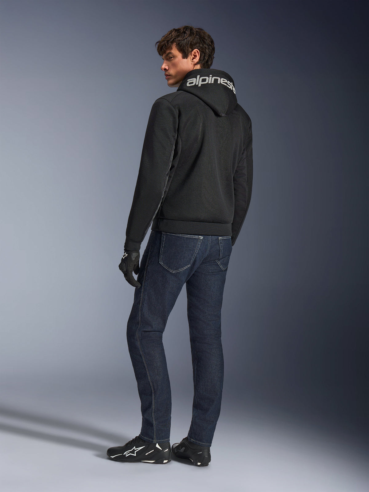 Alpinestars Chrome Superairflow Hoodie, Schwarz, Rückansicht an männlichem Modell, weißes Alpinestars-Branding auf der Kapuze, kombiniert mit dunklen Denim-Jeans und schwarzen Handschuhen, urbanes Motorrad-Design