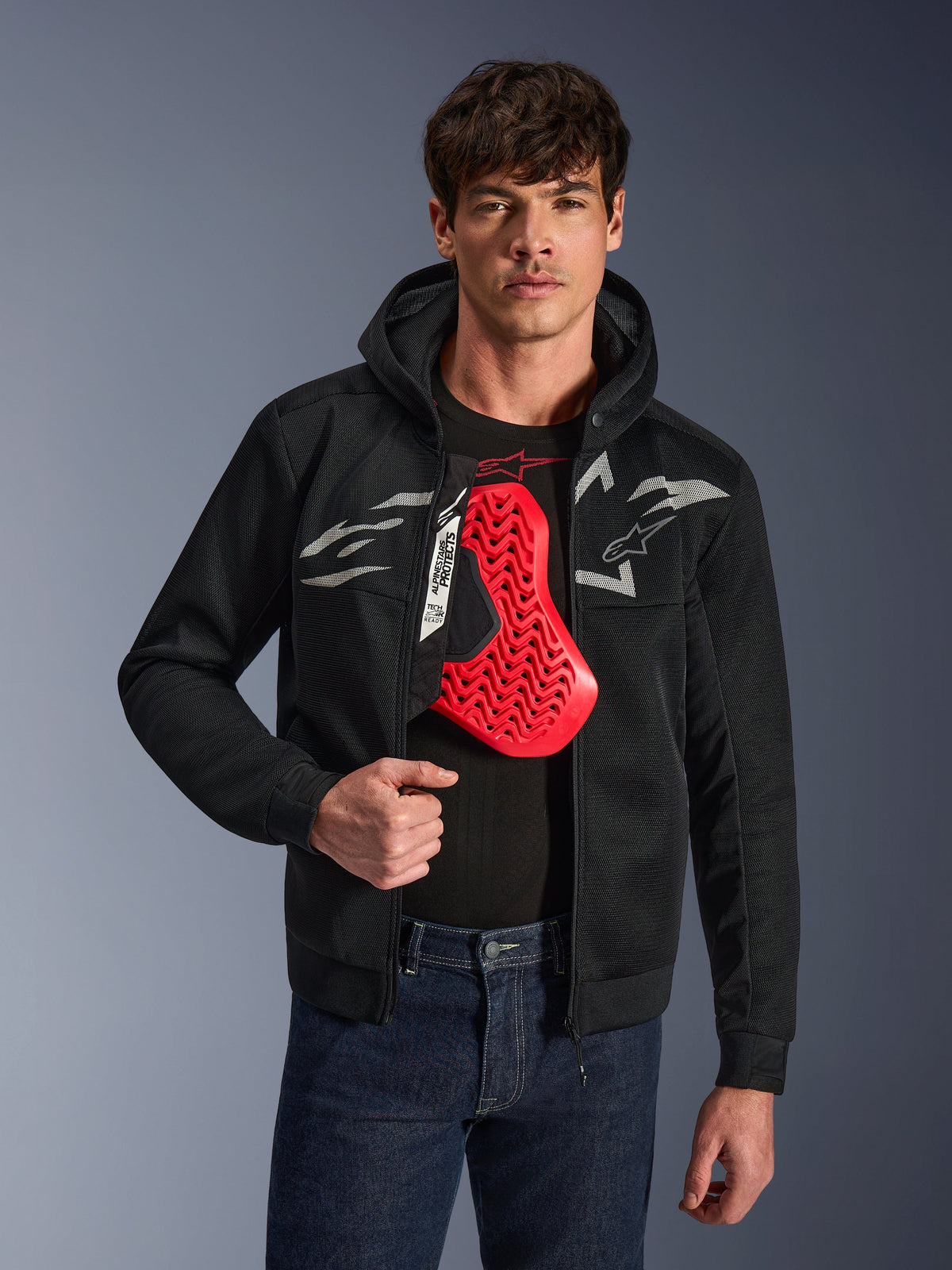 Alpinestars Chrome Superairflow Hoodie in Schwarz, mit weißen und grauen Astars-Logos, getragen von einem männlichen Model mit einem darunter sichtbaren roten Brustprotektor, 3D-Mesh-Textil zur Belüftung, sportliche urbane Motorradbekleidung.
