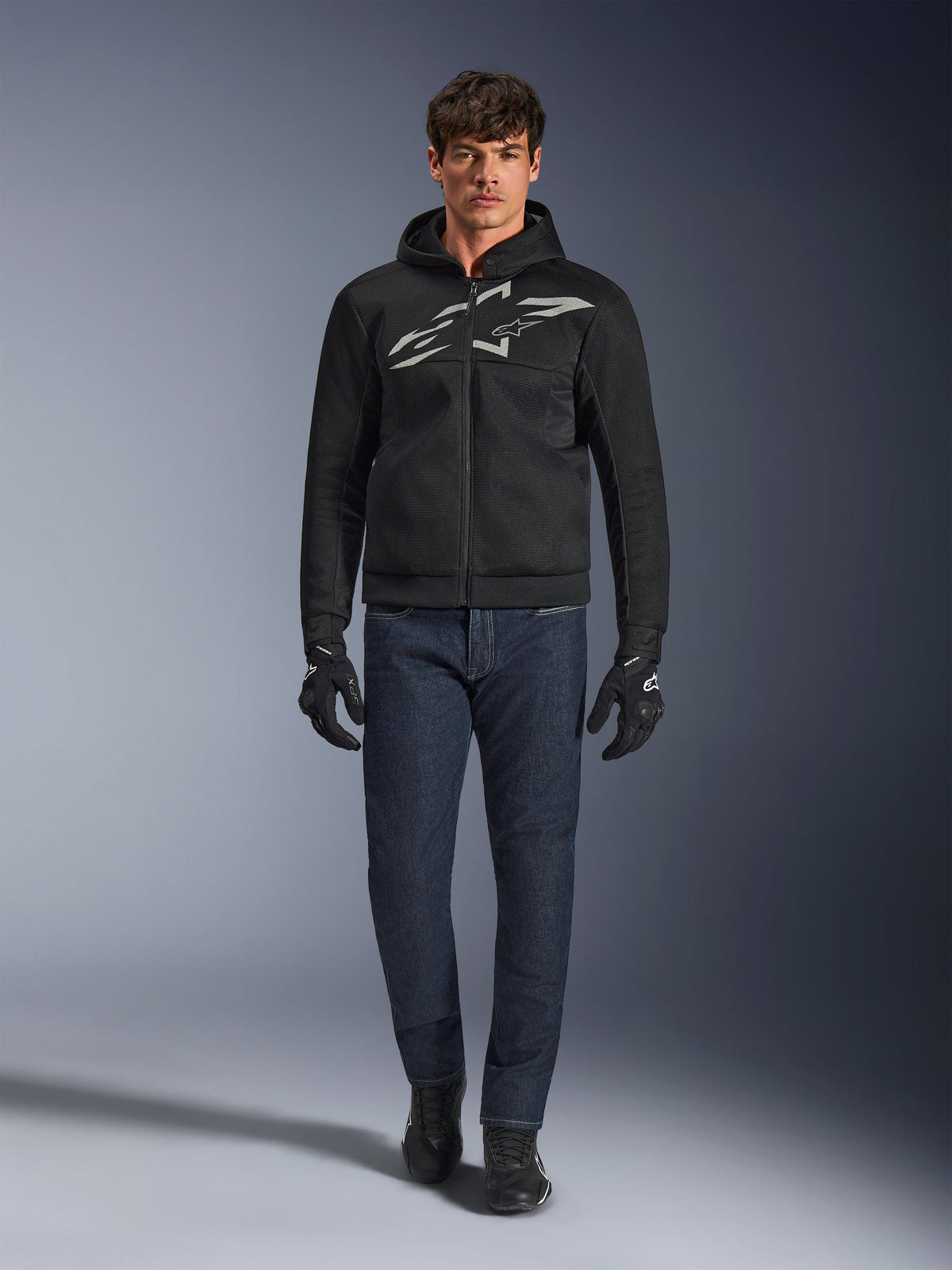 Motorradfahrer trägt den Alpinestars Chrome Superairflow Hoodie in Schwarz und Grau, kombiniert mit dunkelblauer Jeans, schwarzen Motorradhandschuhen und schwarzen Schuhen. Der Hoodie verfügt über ein großes stilisiertes Logo auf der Brust und eine 3D-Mesh-Struktur für Performance bei hohen Temperaturen.