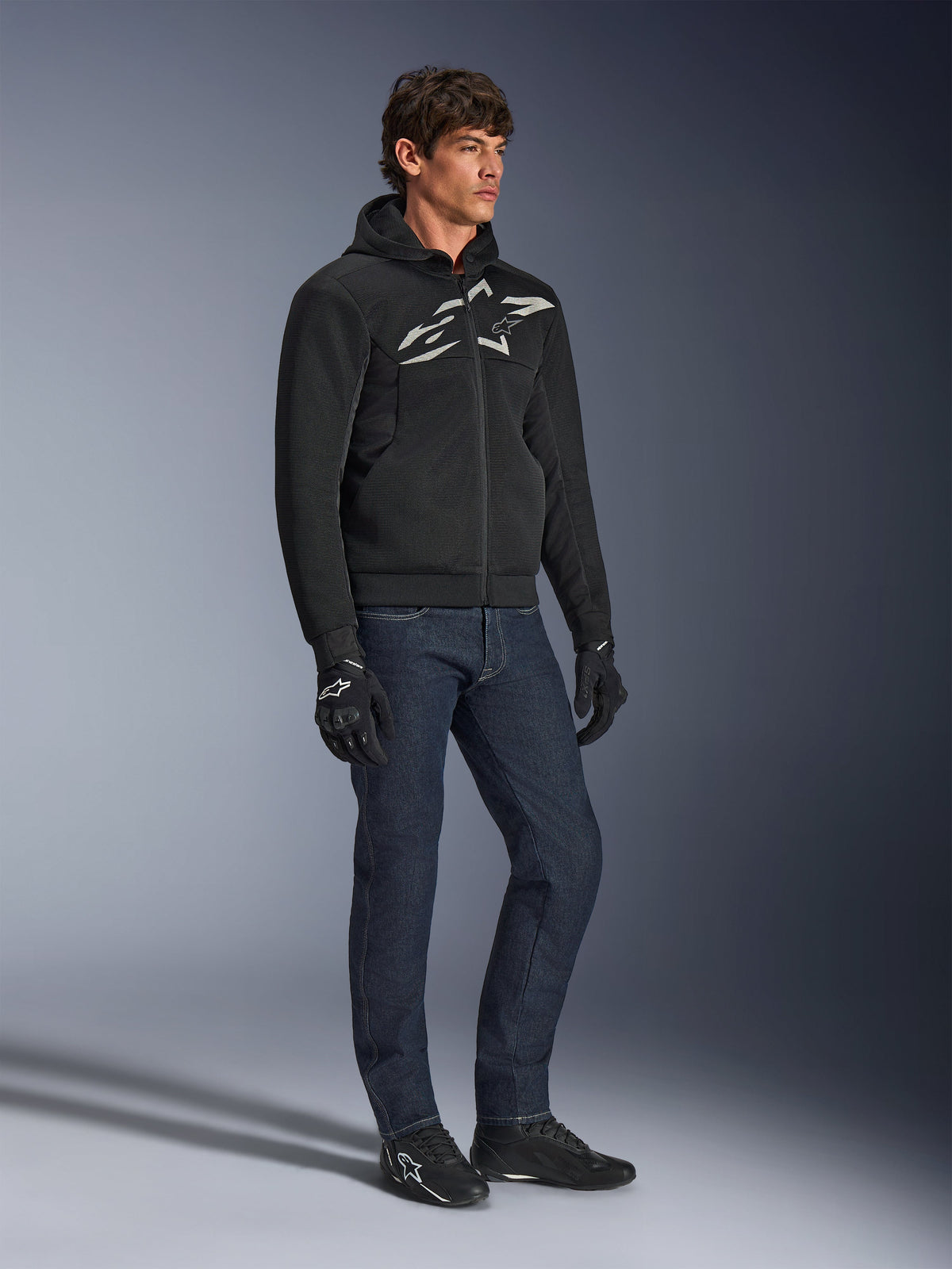Alpinestars Chrome Superairflow Hoodie, schwarz mit weißem und grauem Logo, belüftete 3D-Mesh-Textilkonstruktion, getragen mit dunkelblauen Denim-Jeans und schwarzen Motorradhandschuhen, urbaner sportlicher Fahrstil.