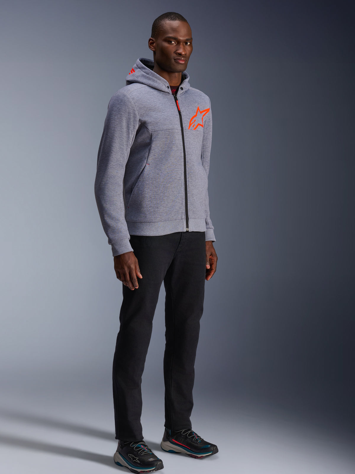Alpinestars Chrome V2 Sport Hoodie, Gray Heather und Red Fluo, getragen von einem männlichen Model mit schwarzer Hose und dunklen Sportschuhen, mit einem leuchtend orangefarbenen Stern-Logo auf der Brust und Kapuze, lässige urbane Motorradbekleidung
