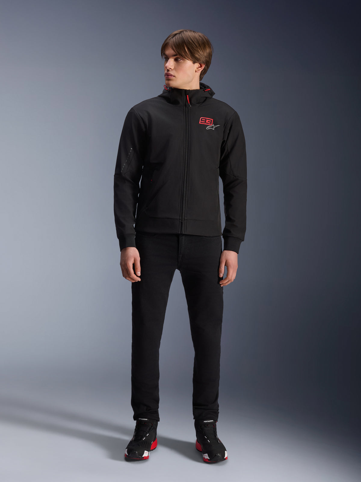 Alpinestars MM93 Radium Tech Hoodie, zwarte urban motorhoodie, voorzien van een rood 93-logo en wit sterlogo op de borst, gedragen door een model met een zwarte broek en bijpassende motorschoenen, slanke winddichte technische shell voor ritten in de stad.