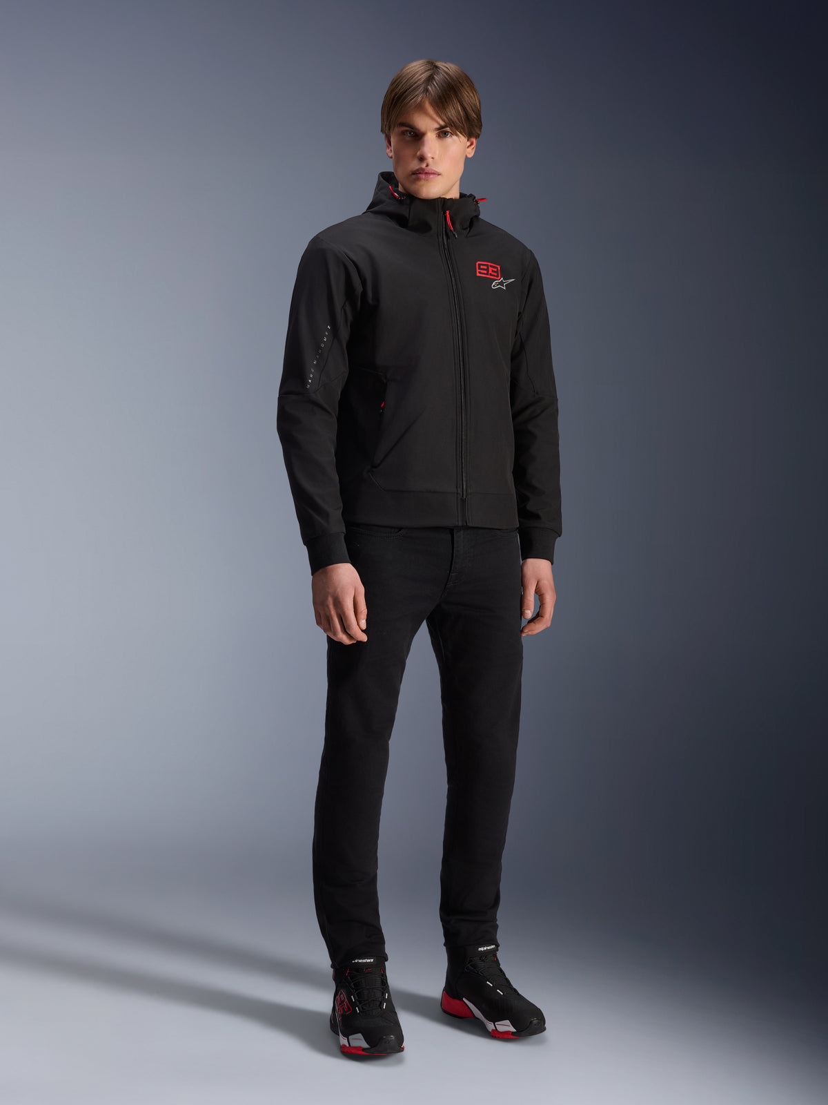 Alpinestars MM93 Radium Tech Hoodie, urban motorjas, zwart met felrode accenten, gedragen door model met zwarte broek en motorschoenen, voorzien van rood '93' logo op de borst en Marc Marquez branding op de mouw, casual technische streetwear voor stedelijk rijden
