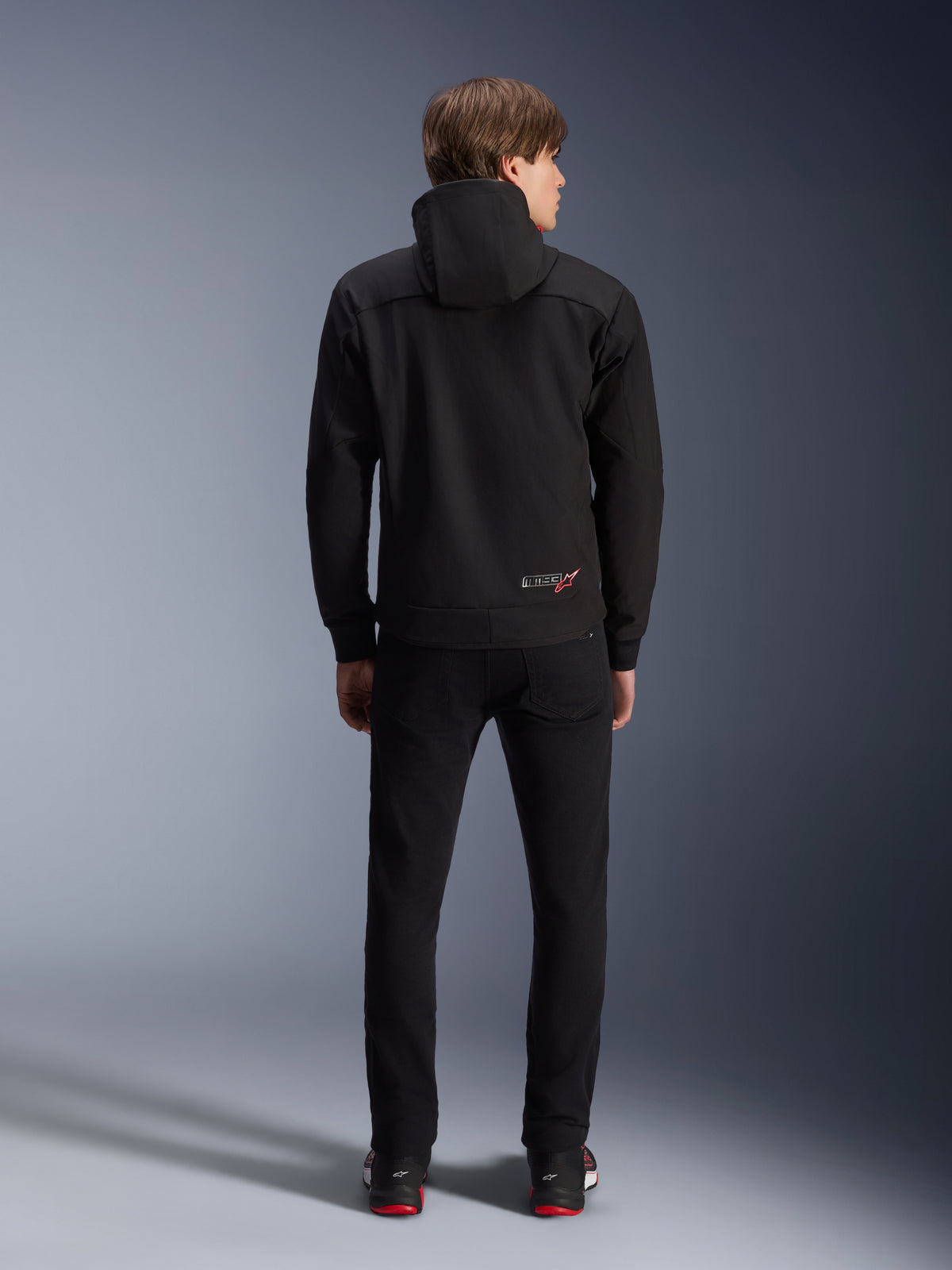 Alpinestars MM93 Radium Tech Hoodie, zwart, achteraanzicht op model, met een klein MM93-logo met rode ster op de onderrug, gecombineerd met een zwarte broek, urban motorkleding