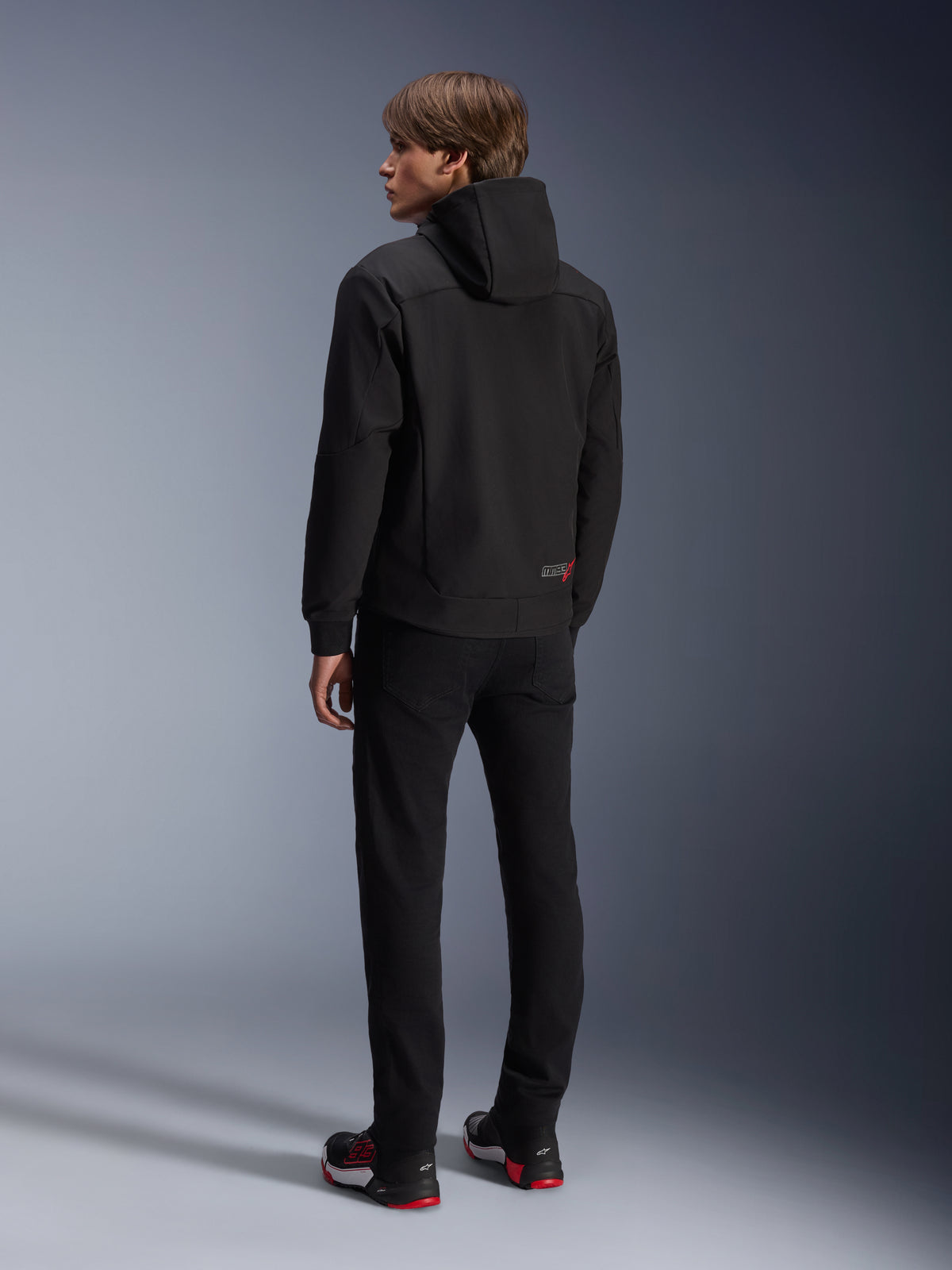 Alpinestars MM93 Radium Tech Hoodie, zwart, urban motorhoodie achteraanzicht, met een klein rood en wit MM93-logo op de onderste zoom, atletische pasvorm met capuchon, gedragen met een zwarte broek en bijpassende rood-zwarte sneakers